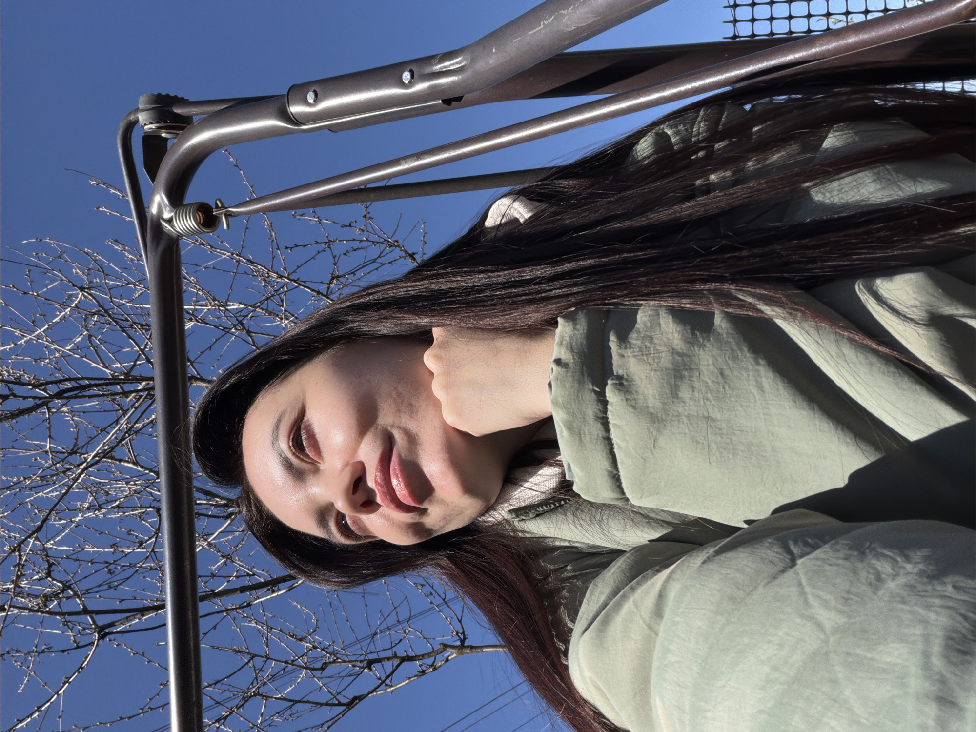 A young woman poses for a selfie outdoors in Chaikovsky, Russia. She is seen from a low angle, looking down at the camera with a slight smile. She has long, dark hair and is wearing a light green, puffy jacket. Above her, a metal structure, possibly part of a swing set or playground equipment, is visible against a clear blue sky. Bare tree branches are also visible in the background.