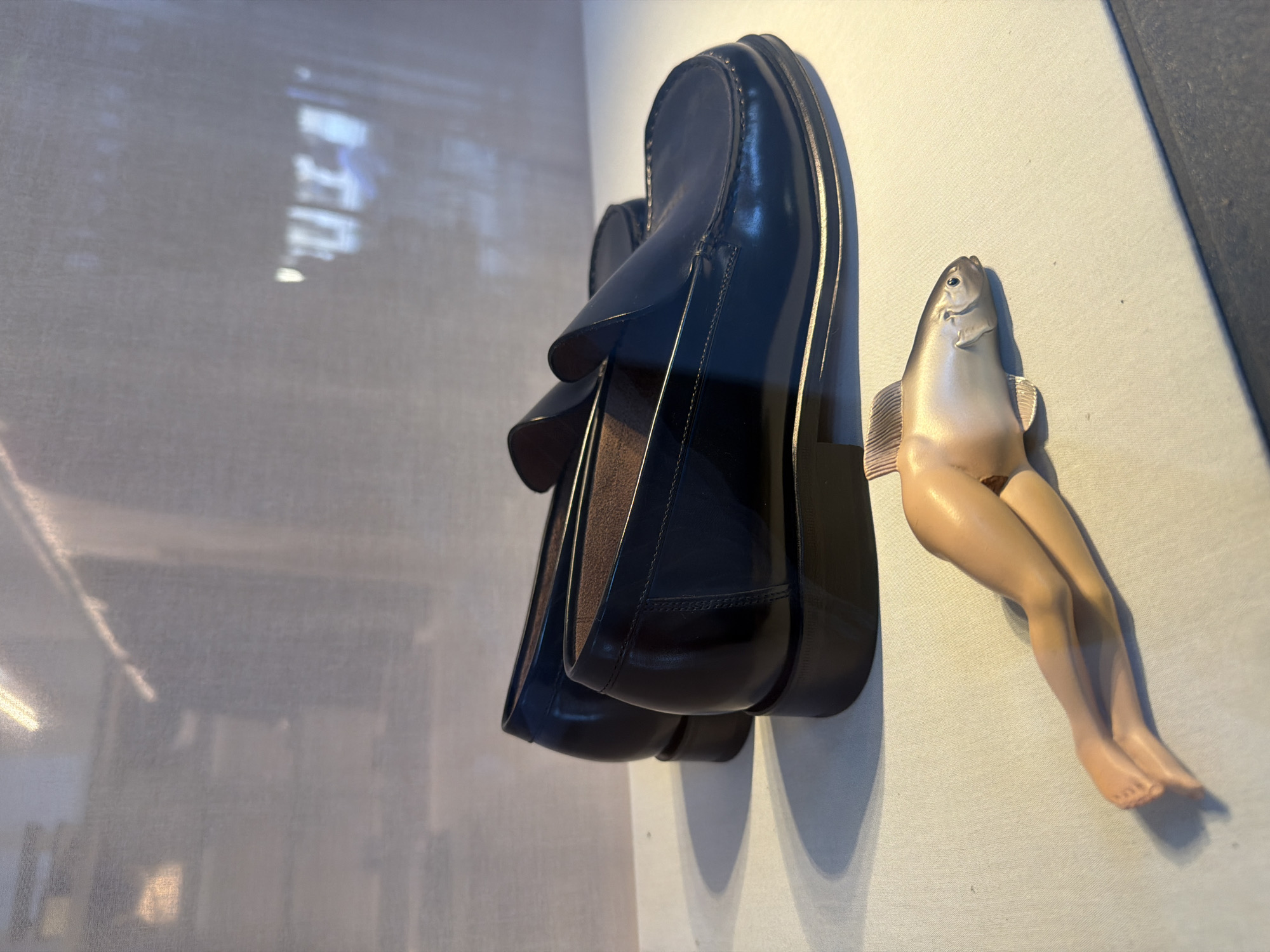 The image presents a surreal juxtaposition of objects against a stark white backdrop. A pair of black leather loafer-style shoes, positioned at an angle, dominates the upper left portion of the frame. To the right of the shoes, a doll-like figure with a fish head and human legs rests. The figure is pale in color and possesses detailed features. The environment appears to be an art gallery or a similar space. The photo's subject and composition suggest a commentary on consumerism, identity, or transformation. I cannot determine the exact location in Paris, France.