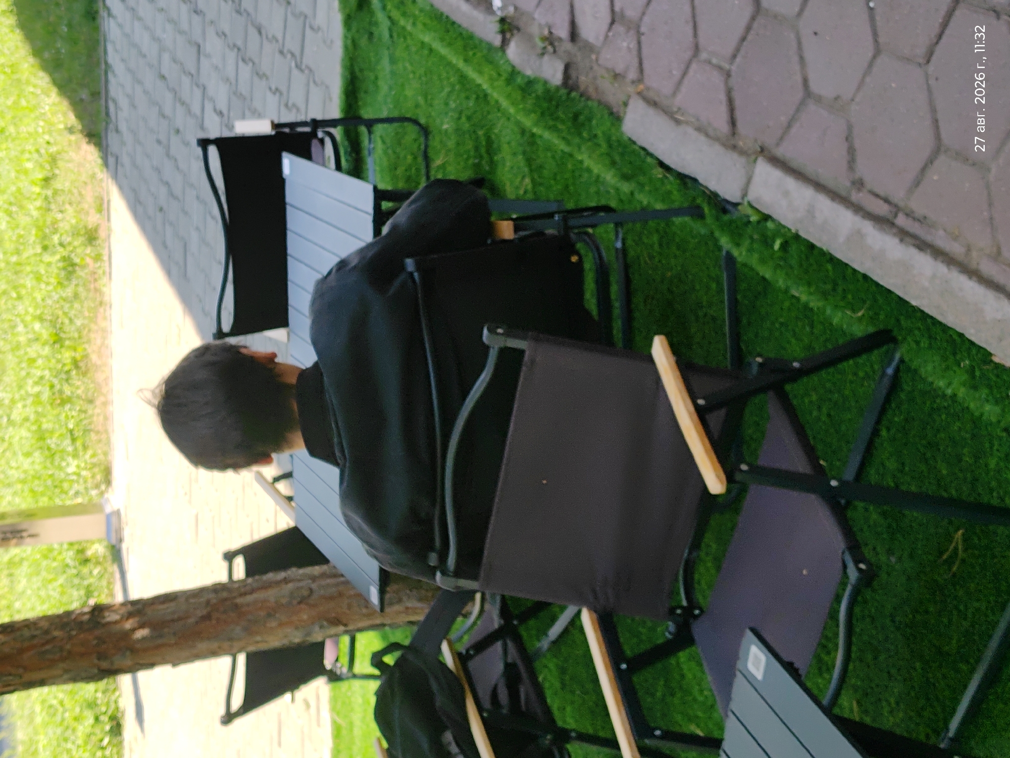 In Astana, Kazakhstan, a person with dark hair, wearing a black jacket, is seated at a black folding table and chair setup in an outdoor area. The setting appears to be a cafe or restaurant with artificial grass flooring and a paved pathway. Additional black folding chairs and tables are visible in the background, along with a tree trunk and a grassy area. The scene is captured from a slightly elevated, rear-facing perspective.