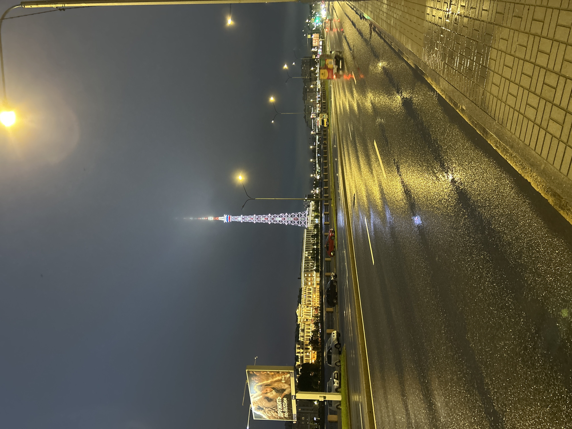 An exterior, eye-level, medium shot captures a city street at night. The street is wet, reflecting the yellow glow of streetlights and the illuminated spire of a broadcast tower in the distance. The tower is tall and slender, with a lattice structure adorned with red and white lights. Several cars are visible on the road, some moving and others parked. On the left side of the street, a large billboard displays an advertisement. The sidewalk is paved with yellow bricks and is also wet. The sky is dark and overcast, with a streetlamp visible in the upper right corner. The overall impression is one of a damp, urban nighttime scene.
