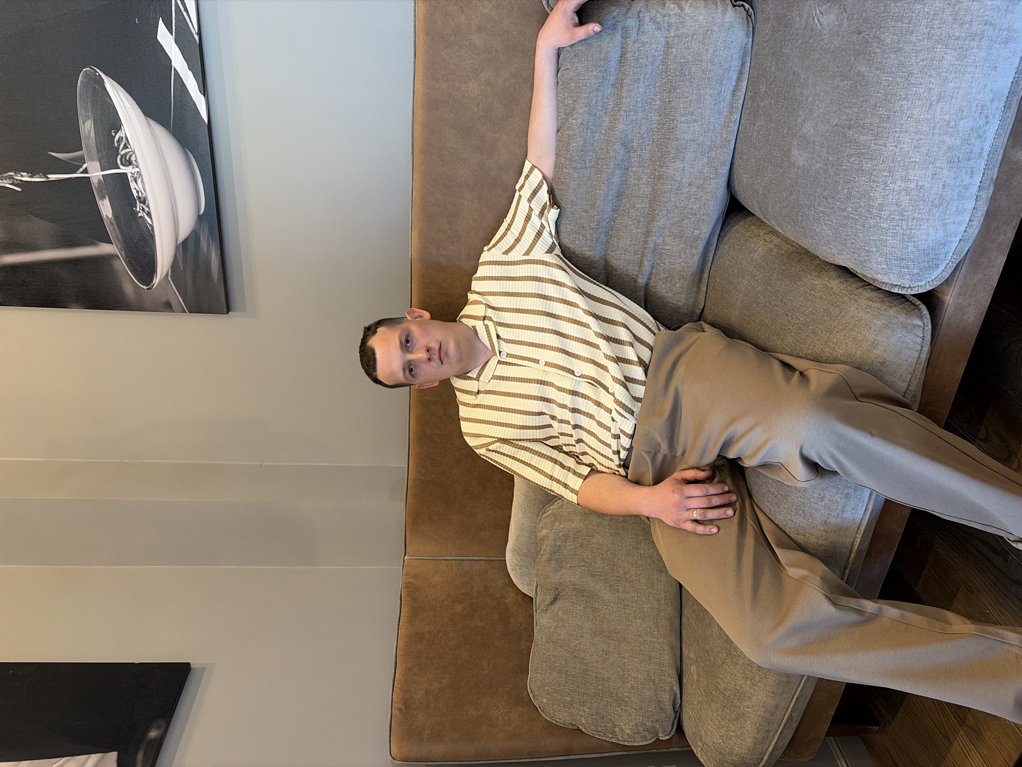 A man sits on a brown and grey couch in a room with grey walls. A black and white picture hangs on the wall. The man wears a striped, short-sleeved button-up shirt and khaki pants. He has short hair and is looking at the camera. The couch has multiple cushions. The location is potentially indoors, given the furniture and wall decor, but is not conclusively identified in Yekaterinburg, Russia.