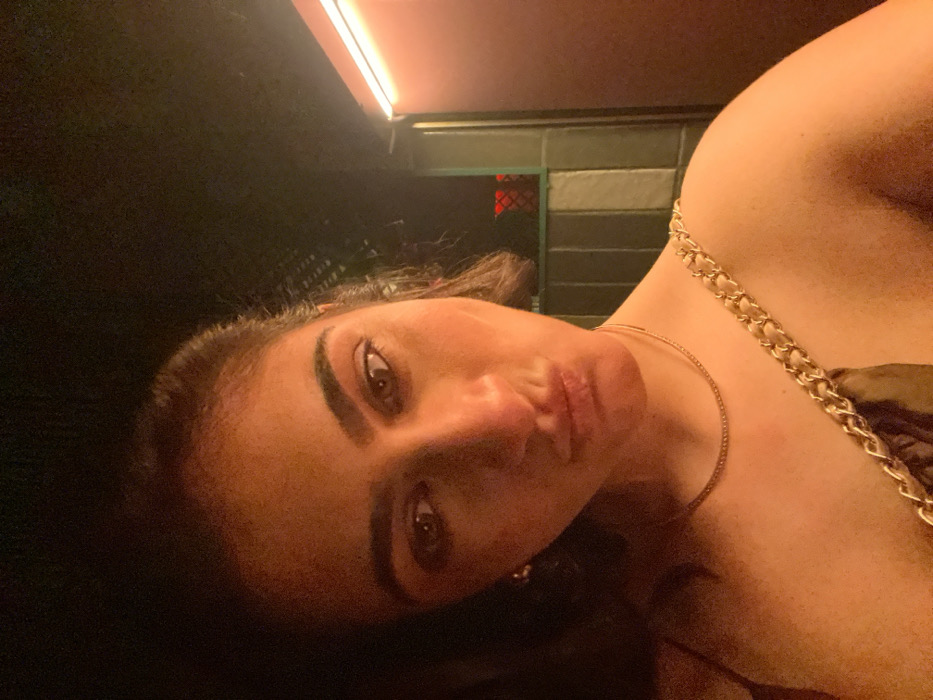 A female individual is depicted in a chest-up, slightly off-center portrait shot, facing directly towards the camera. She has dark hair, brown eyes, and is making a puckered "duck face" expression. Her skin, particularly her face and exposed shoulder, is illuminated by a dominant warm, reddish-orange artificial light source, casting soft shadows. She is wearing a dark top, with a thin strap visible on her left shoulder and a distinctive wide strap composed of interlocking gold chain links across her right shoulder. A thin gold necklace is worn around her neck, and a small, circular earring is visible on her left ear.
The background is dark and largely out of focus. To the right of the subject, a vertical strip of bright red-orange light, likely neon or LED, is visible, illuminating an adjacent dark panel or wall section. Further back and to the right, behind the subject's shoulder, a dark, possibly gridded or barred structure is faintly visible, subtly illuminated by a muted red light from within or beyond it. The environment suggests an indoor or semi-enclosed urban setting, consistent with a bar, club, or similar nightlife venue, located in Madrid, Spain.