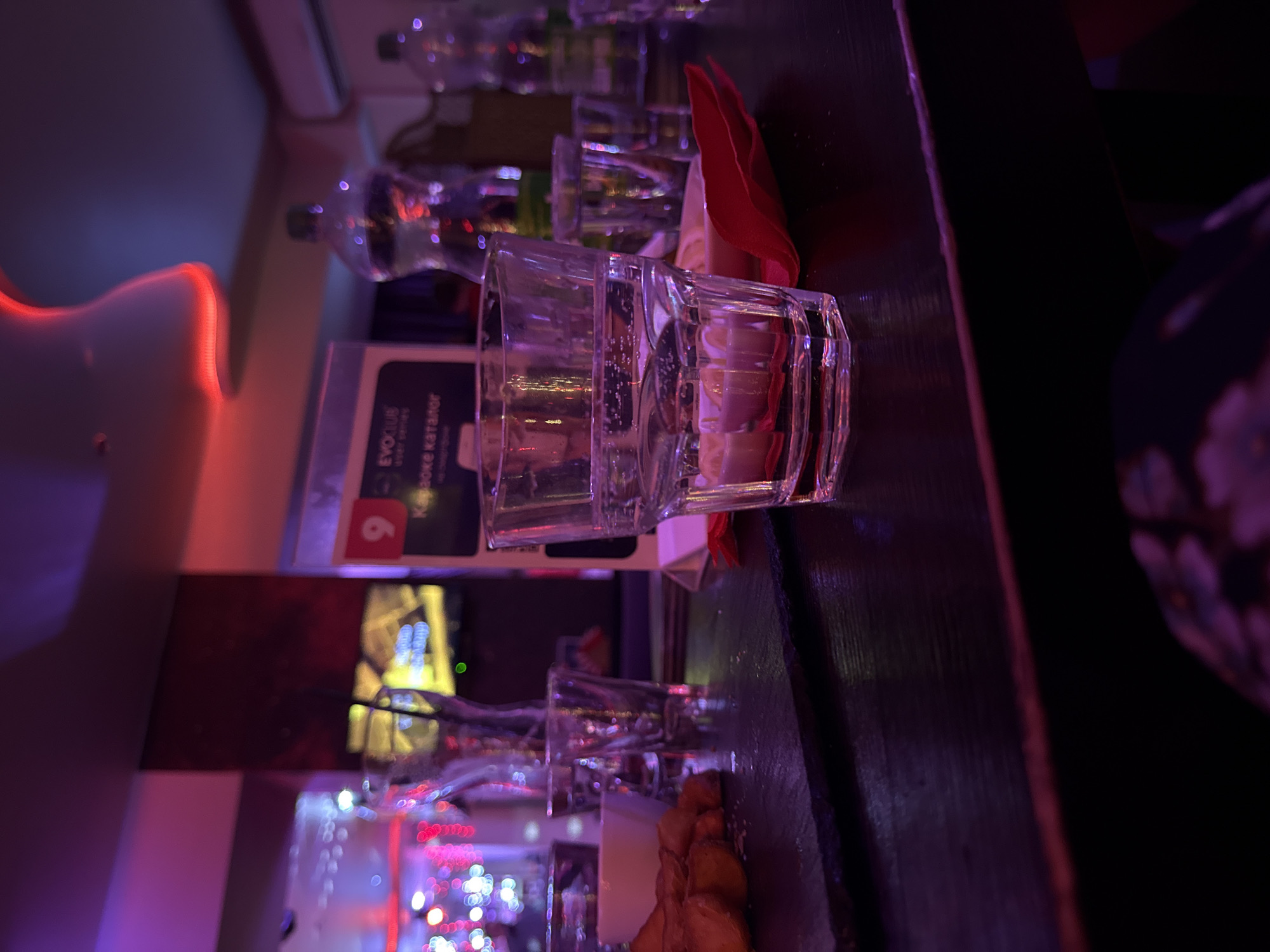 The image depicts a bar scene, possibly in Votkinsk, Russia, featuring a row of shot glasses and water bottles lined up on a dark-colored countertop. The foremost shot glass is prominent, capturing the light and revealing some liquid inside, potentially vodka or a similar spirit. A folded red napkin is placed near the shot glasses. Behind the glasses and bottles is an advertisement card, likely for a karaoke system from the brand "EVOKLUB."
In the background, partially blurred, there are lights suggesting a nightlife ambiance, with festive decorations. The colors of the environment are dominated by reds, blues, and purples, indicating the presence of artificial lighting typical of bars and nightclubs.
The presence of water bottles suggests that the patrons are being served refreshments alongside the alcoholic beverages. An individual wearing a garment with a floral pattern is partly visible in the lower-right corner, implying that other people are present in the scene.