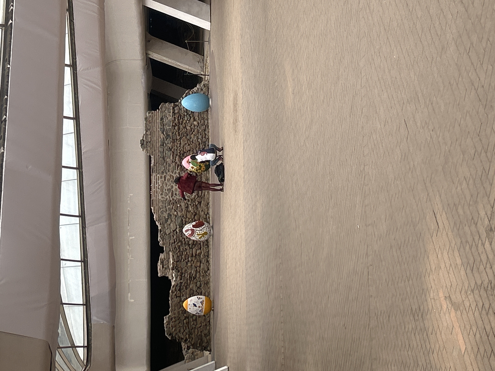 A slightly rotated, eye-level, outdoor shot reveals an architectural space with large, decorated Easter eggs along a stony structure. The location is likely Sofia, Bulgaria, given the Easter egg theme.
The left portion of the image features a white structure with long vertical window panes. Adjacent to it, a weathered stony or concrete wall, possibly a decorative or historical feature, runs vertically down the frame.
Four large, decorated Easter eggs are placed along the stony structure at varying heights. The designs on the eggs are not clearly discernible.
Three people are visible from an aerial perspective, positioned near the middle of the frame. One person is wearing a red outfit.
To the right of the stony structure, a broad expanse of grey brickwork extends into the background, hinting at a large public space or square. The pavers are laid in a uniform, textured pattern.