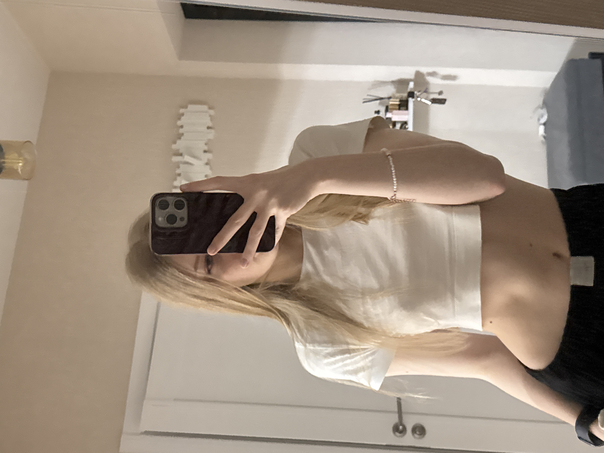 A young woman is captured in a mirror selfie, appearing to be taken in an indoor setting, likely a bedroom or dressing area in Perm, Russia. She holds a dark-colored smartphone, possibly an iPhone due to its distinct multiple camera lenses, in her right hand, partially obscuring her face. She has light-colored hair, which cascades over her shoulders and chest. She is wearing a white, short-sleeved, cropped t-shirt that reveals her midriff, and black bottoms, which could be trousers or a skirt. A delicate bracelet adorns her left wrist, and a dark-colored watch or fitness tracker is visible on her right wrist.
The reflection in the mirror reveals elements of the room behind her. To the left, a gold-colored object, possibly a decorative lamp, is mounted on the wall. A unique white, textured object, resembling stacked vertebrae or a modular shelf, is also affixed to the wall. Further back, a collection of items, including bottles and containers, can be seen on what appears to be a shelf or dresser. A light-colored door or cabinet is partially visible below the phone. In the deeper background reflection, a bed or sofa with a blue covering can be vaguely discerned. The lighting suggests an interior setting, likely under artificial light. There is no visible text in the image.