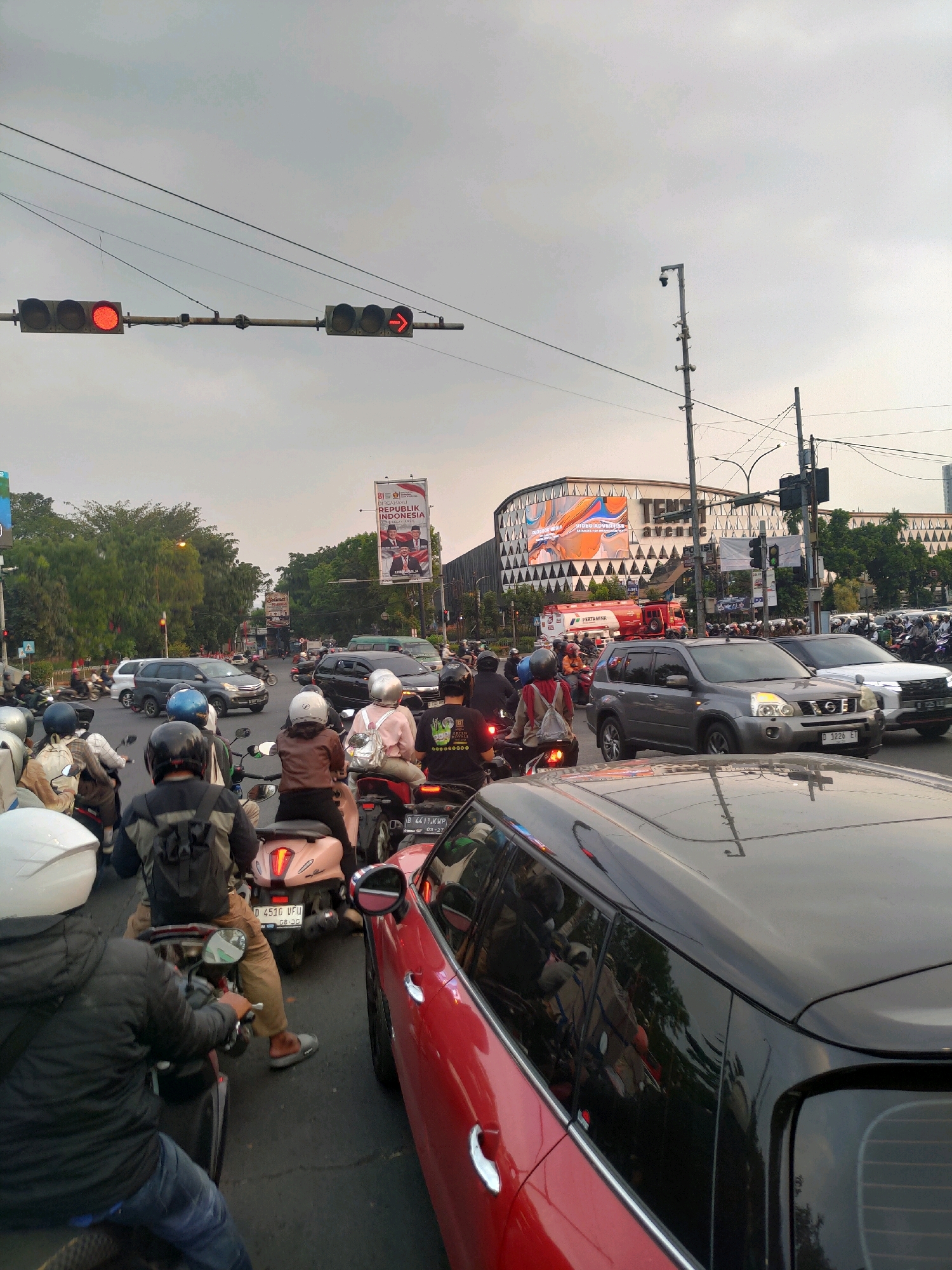 The image captures a busy intersection in Bandung City, Indonesia, during what appears to be traffic congestion. A red traffic light is illuminated on the left, indicating a stop. To its right, a traffic signal displays a red arrow pointing right, also signaling a stop for turning traffic. The sky is overcast, suggesting a cloudy day.
Numerous vehicles, primarily motorcycles, are stopped on the road, with some drivers and passengers visible. People are wearing helmets, indicating adherence to traffic safety regulations. A red and black car is prominently positioned in the foreground, with its reflection visible in the tinted windows of another vehicle. In the background, a modern building with large illuminated advertisements stands out, and a red fire truck with "PERTAMINA" branding is also present. Billboards displaying official imagery are visible, suggesting a public or government-related event or campaign. The overall scene depicts a typical urban traffic scenario with a mix of vehicles and infrastructure.
