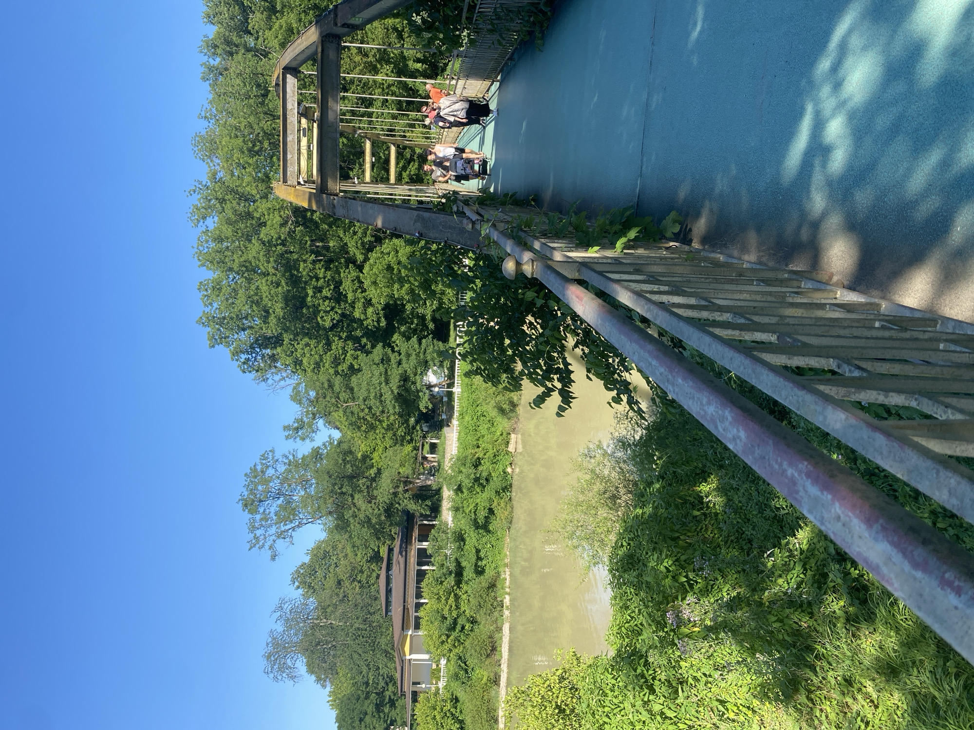 A view from a pedestrian bridge in Goryachiy Klyuch, Russia, looks out over a muddy river and lush green trees. The bridge itself has a light blue pathway and a weathered metal railing. On the bridge, several people are walking, some with a baby stroller. In the distance, across the river, a light-colored building with a dark roof is visible, surrounded by trees. The sky is clear and blue.