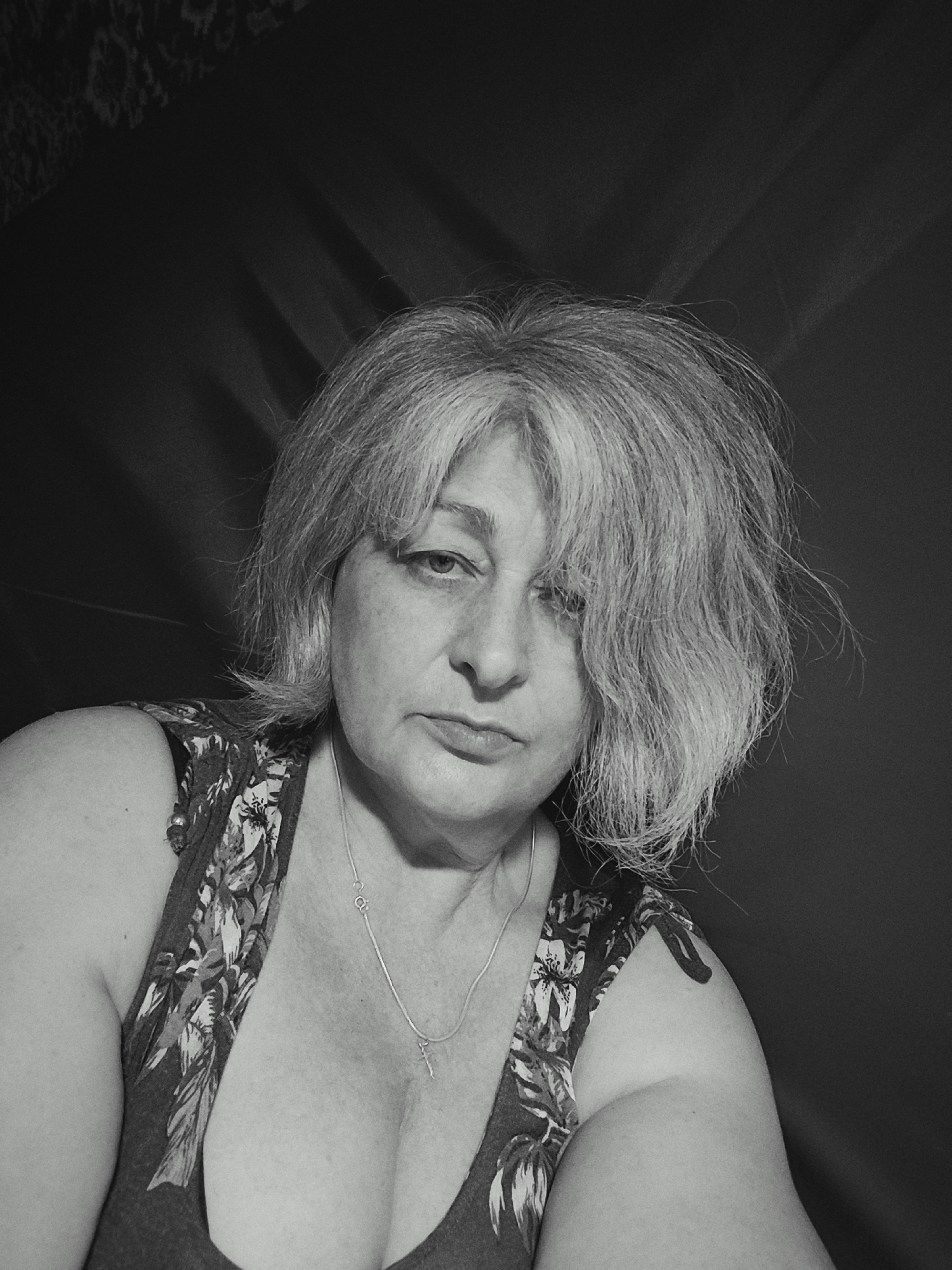 A black and white, medium close-up shot shows a middle-aged woman with light-colored, tousled hair looking directly at the camera. She has a neutral expression with her mouth slightly downturned. The woman is wearing a sleeveless top with a floral pattern and a silver necklace. She is positioned in front of a dark, textured background that appears to be a draped fabric. The lighting casts shadows on the background, suggesting it is coming from the side and slightly from above. There is no identifiable location in Krasnodar, Russia, in this image.