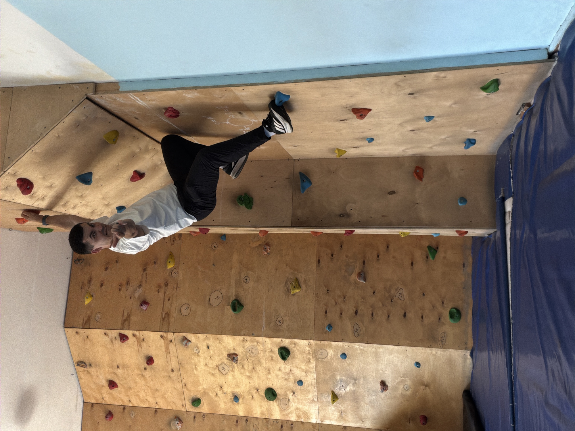 A man is climbing an indoor climbing wall. He is wearing a white t-shirt and black pants. His left foot is on a blue climbing hold near the top of the wall. He is looking at the camera and pointing with his right hand. The climbing wall is constructed of plywood and has various colored climbing holds attached to it. The wall is located indoors, with a light blue ceiling visible above the wall. There is a blue protective tarp on the right side of the wall. The location is possibly Izhevsk, Russia.