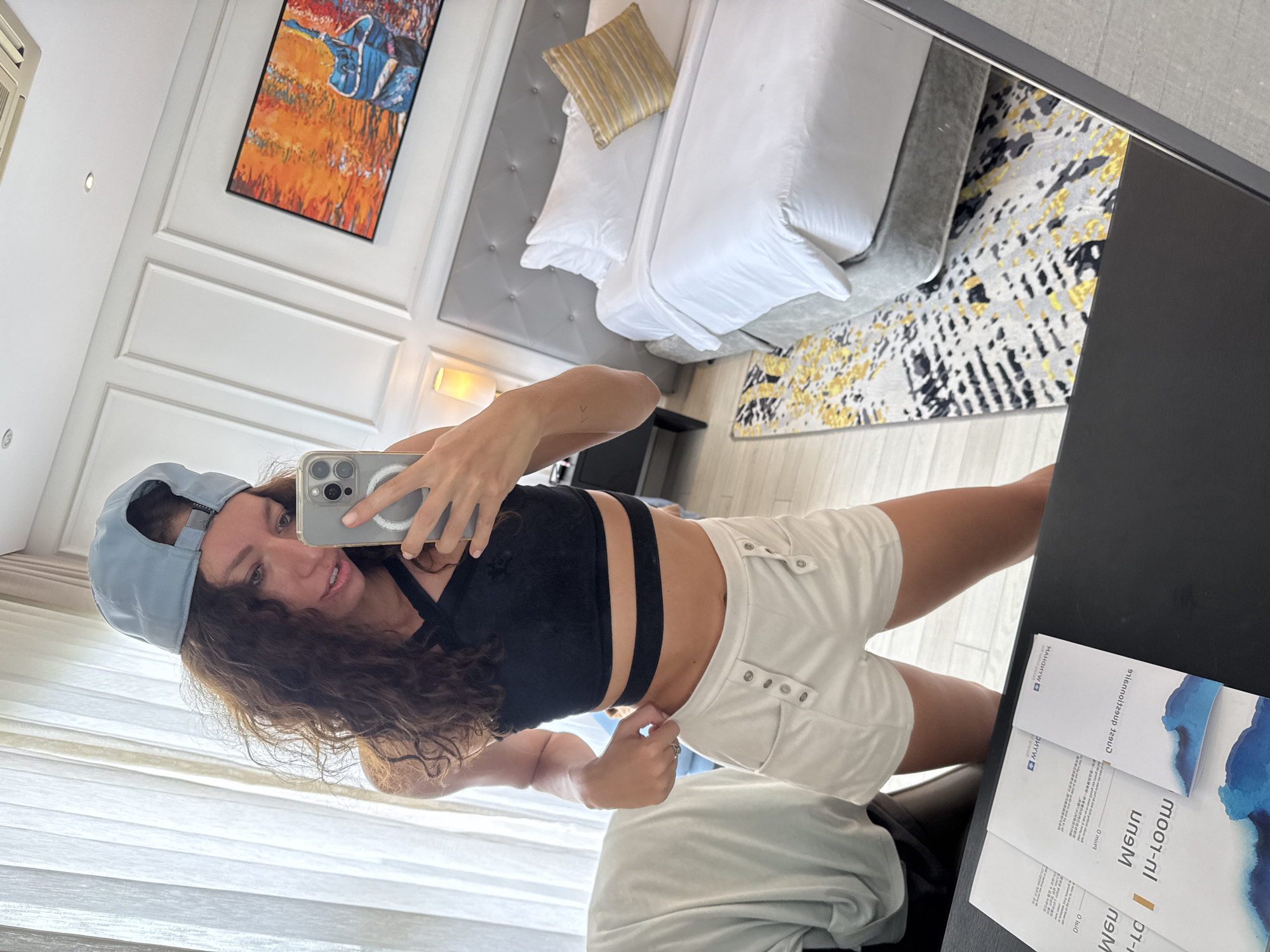 A woman with curly brown hair is taking a selfie in a hotel room in Đà Nẵng, Vietnam. She is wearing a blue baseball cap, a black sports bra, and white shorts. She is holding a silver iPhone in her right hand. Behind her is a bed with white linens and a grey headboard. A painting hangs on the wall. A black desk is visible in the foreground, with brochures on top. The room has light-colored walls and curtains.