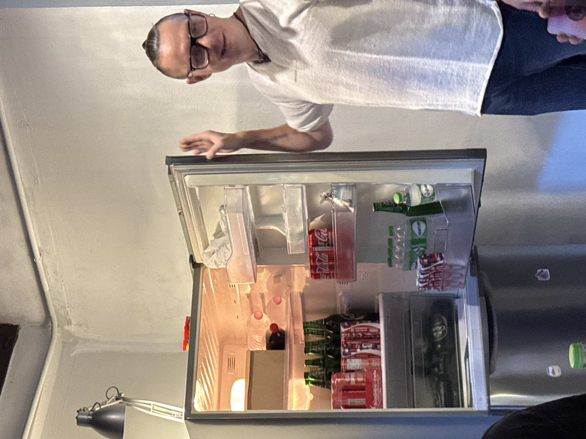 A man, wearing glasses and a white t-shirt, is captured in an upside-down, close-up shot, reaching into an open, illuminated refrigerator. The refrigerator is filled with various beverages, including bottles of water and cans of Coca-Cola. In the foreground, there are several rows of green glass bottles with white labels, and some red and white cans stacked on shelves. The man's hand is extended into the refrigerator, seemingly in the process of selecting an item. The scene appears to be indoors, possibly a home or a small establishment in Chiang Mai, Thailand, given the provided location context. The lighting within the refrigerator is bright, illuminating the contents and casting a warm glow. The man's expression is not clearly visible due to the angle and resolution of the image, but his posture suggests casual interaction with the refrigerator's contents. The background consists of a plain, light-colored wall and a portion of what looks like another appliance, possibly a washing machine or a larger refrigerator. The overall impression is a candid moment of someone accessing drinks from a well-stocked fridge.