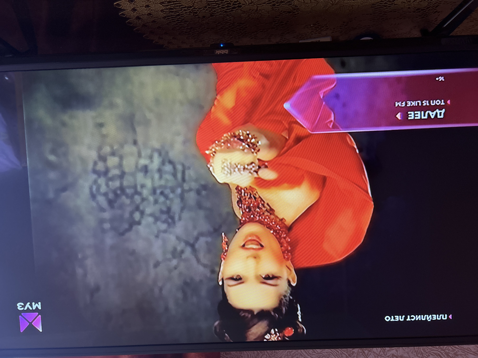 The image displays a television screen showing a music channel interface. The channel logo, a stylized purple "MY3," is visible in the upper right corner. On the left side of the screen, text indicates "ПЛЕЙЛИСТ ЛЕТО" which translates to "SUMMER PLAYLIST." Below this, a purple banner reads "ДАЛЕЕ" ("NEXT") and "ТОП 15 LIKE FM" ("TOP 15 LIKE FM"), with "16+" indicating age restriction.
The central subject of the broadcast is a woman in a vibrant red dress, adorned with elaborate jewelry including a chunky, multi-layered red necklace and large, dangling earrings. Her hair is styled in an updo, and she is wearing dramatic eye makeup. Her expression is animated, with her mouth open as if singing or speaking. Her hands are positioned forward, also decorated with ornate rings.
The background of the video appears to be a textured, dark gray or black surface, possibly a studio backdrop. The television itself is a flat-screen model, with a visible "bbk" logo on the bottom bezel. The screen is placed on a surface covered with a patterned tablecloth. The image is taken from a slightly elevated angle, suggesting the television is positioned in a room. The overall scene captures a moment from a music video or program broadcast on a Russian music channel. The location is not specified in the image, and therefore, its presence in Gelendzhik, Russia cannot be confirmed.