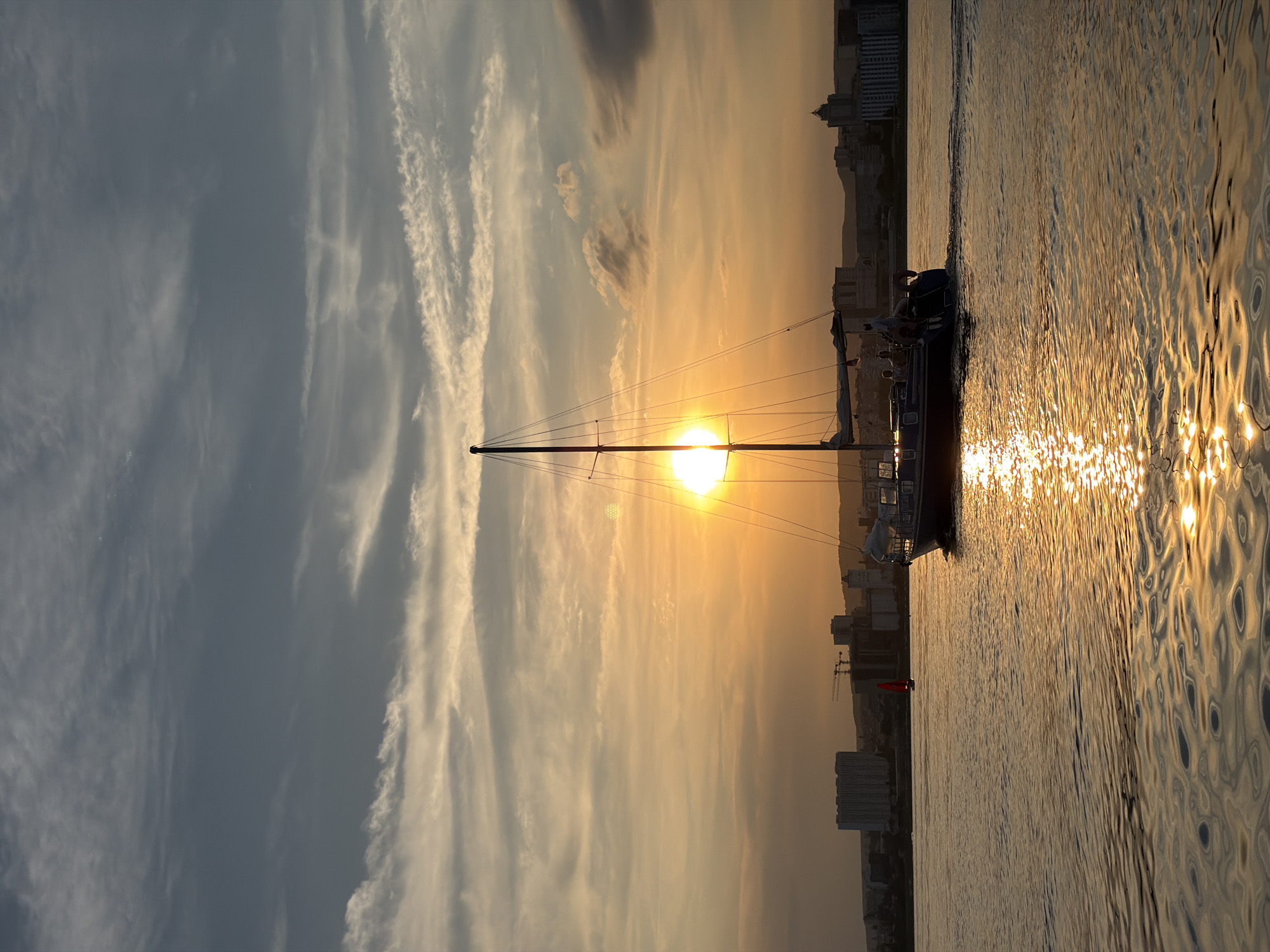 The image captures a serene sunset over the Volga River in Saratov, Russia. The sun, a bright orange orb, is positioned just above the horizon, casting a warm, golden glow across the water and the sky. Wispy clouds, illuminated by the sun's rays, stretch across the twilight sky, transitioning from a pale blue to hues of orange and gold.
In the foreground, a sailboat glides across the rippling water. Its dark hull is silhouetted against the vibrant sunset, and its mast stands tall, adorned with rigging. Several figures are visible on the deck of the sailboat, enjoying the tranquil evening. The reflection of the setting sun shimmers on the water's surface, creating a dazzling display of light.
In the background, the Saratov skyline emerges. A cluster of buildings, including a large, modern structure and some industrial cranes, dots the distant shore. The city lights are beginning to twinkle, adding to the picturesque scene. A smaller red sailboat can be seen in the mid-distance, adding a touch of color to the water. The overall atmosphere is one of peace and tranquility, a perfect moment captured at the end of a day.