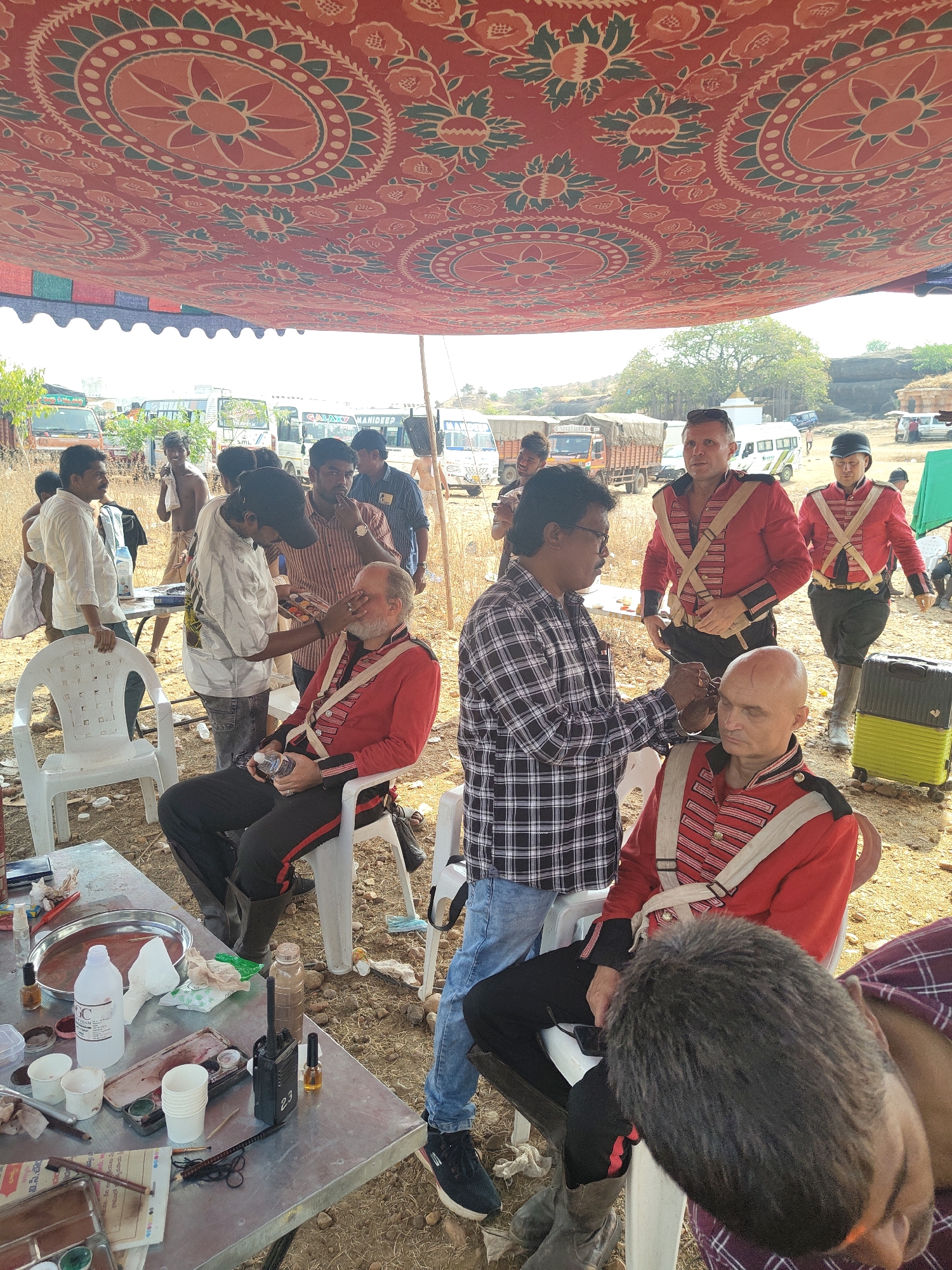 The image depicts a scene in Vamasamudram, India, possibly a film set. Several people are gathered outdoors, under a fabric awning. Two men dressed in red military-style uniforms with white cross-straps are seated on white plastic chairs, receiving makeup touch-ups from two other men. One man in a red uniform stands nearby while another one walks away with a suitcase. A group of onlookers surrounds the scene.
In the background are several vehicles, including buses and trucks. There is a table with makeup supplies. The ground is dusty and exposed with small rocks and dried grass. A temple or other structure sits on a rock formation in the distance.