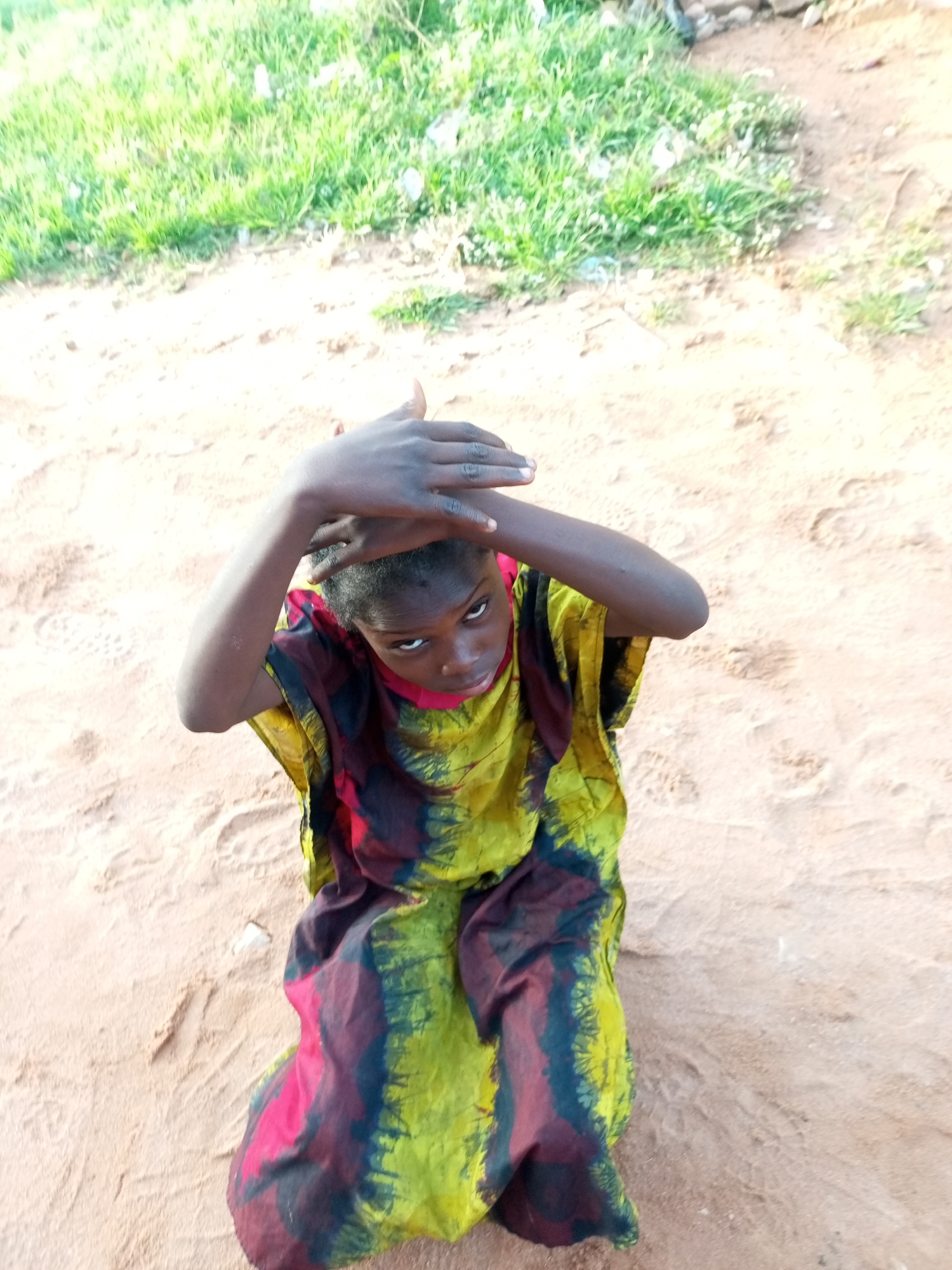 A young person, identified as female by their clothing, is kneeling on sandy ground, looking upward with hands raised to their head. They are wearing a colorful, tie-dyed garment in shades of yellow, green, red, and black. The background consists of sandy ground and a patch of green grass. The scene appears to be outdoors, possibly in a rural or undeveloped area of Tempure, Nigeria. The lighting suggests it is daytime.