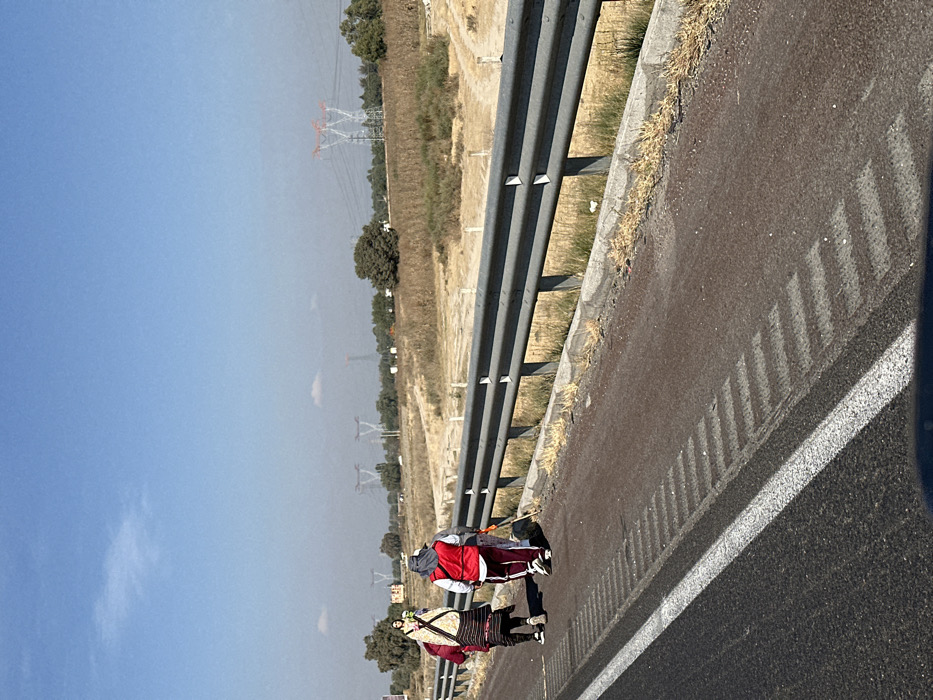 Two people are walking along the shoulder of a highway. A guardrail runs parallel to them, separating the road from a field dotted with power lines. The sky is bright blue, but a haze obscures the distant landscape. The road has painted white lines to mark the lanes. One person wears a red jacket, while the other wears a patterned garment. Their shadows stretch onto the road.