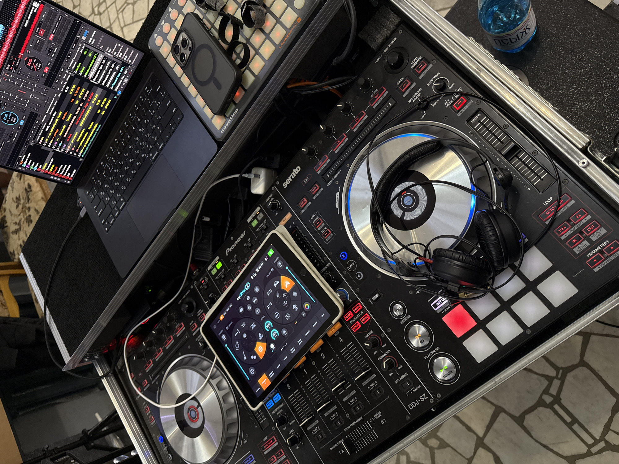 The image shows a DJ setup in what appears to be a domestic setting, possibly in the city of Zavod Mikhailovskii, Russia, as indicated by the provided context. The setup includes a Pioneer DDJ-SZ DJ controller, a laptop running DJ software, an iPad displaying DJ software, a Novation Launchpad S, and a smartphone with a magnetic case. A pair of headphones rests on one of the DJ controller's platters. The equipment is housed within a black flight case. The background suggests a patterned floor and furniture, contributing to the impression of an indoor, possibly residential, environment. There are no people visible in the image. The lighting is artificial, likely from overhead sources.