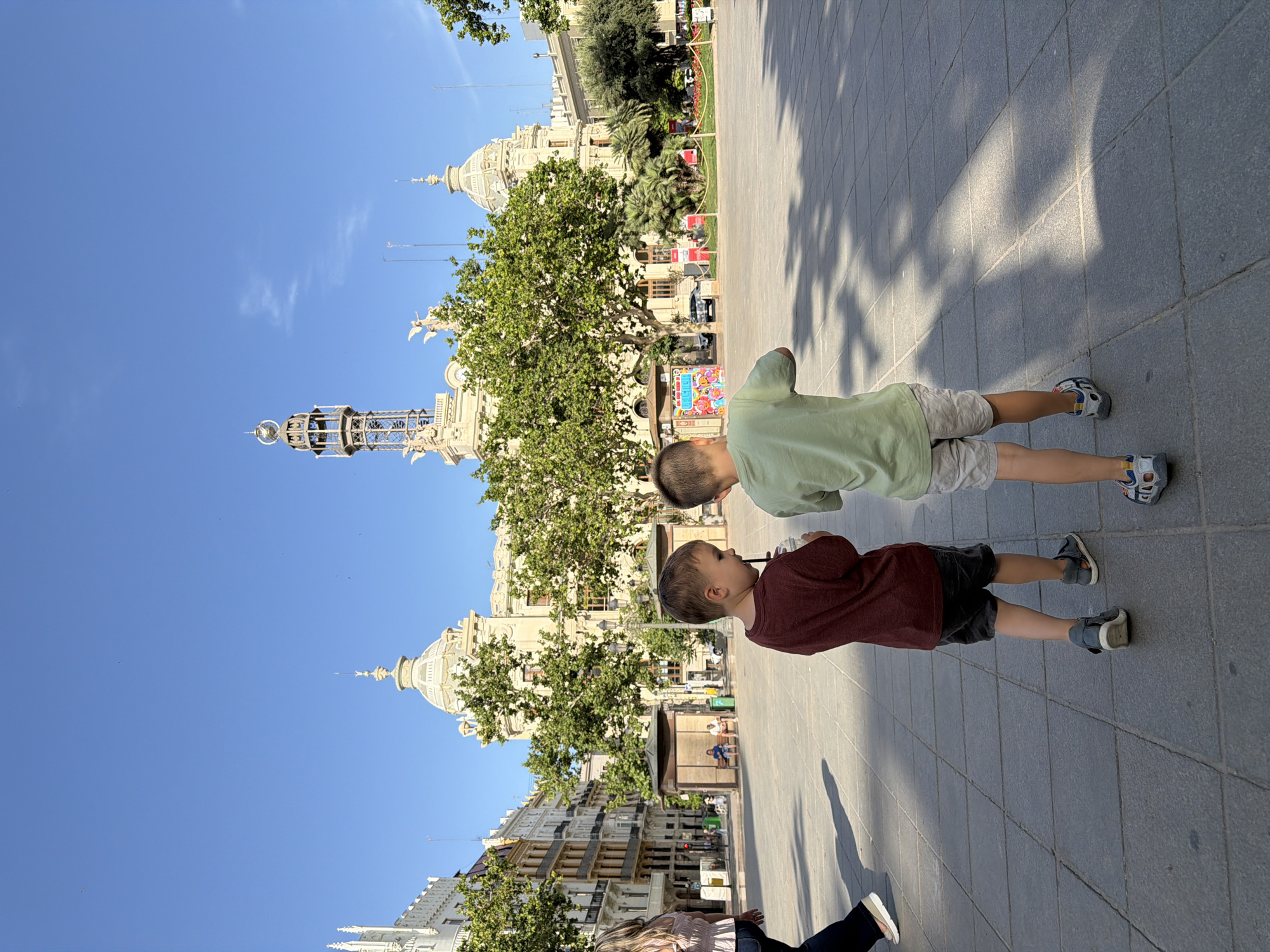 Two young boys walk away from the camera on a paved plaza in front of a large, ornate building. The boy on the left wears a maroon t-shirt and dark shorts, holding a drink with a straw. The boy on the right wears a light green t-shirt and patterned shorts. Both boys wear sneakers. In the background, the building features domes and a tower, with lush green trees in front of it. Sunlight casts shadows on the plaza. A woman in a pink top and dark pants walks to the left in the foreground. The sky is clear and blue.