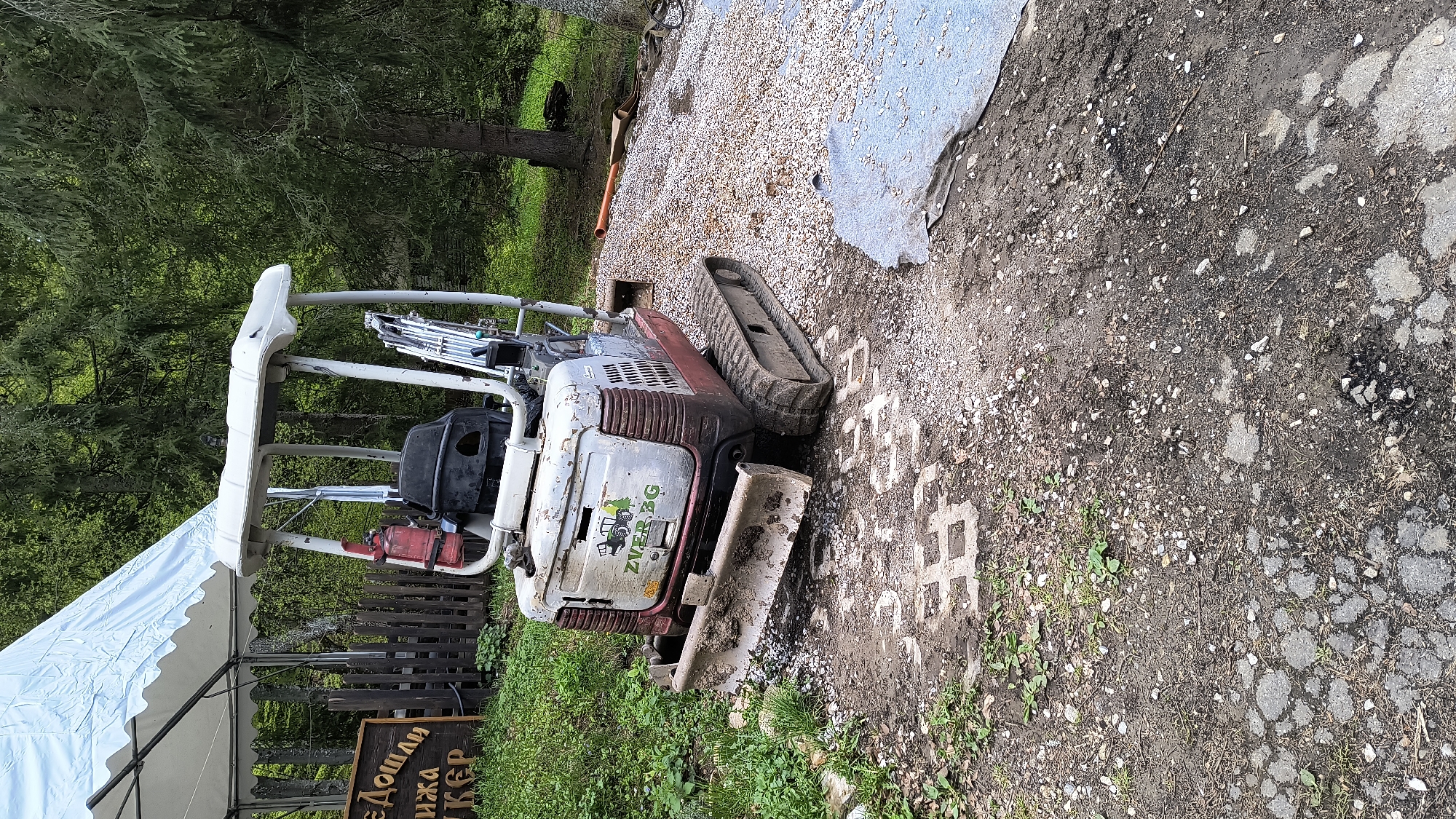 The image shows a mini excavator, likely a Kubota model based on its design, parked on an uneven surface covered with gravel and broken asphalt. The surrounding area is partially paved with interlocking concrete pavers. Patches of green grass and vegetation are visible, indicating an outdoor environment.
To the left, a white tent structure is partially visible, next to a wooden fence and a sign with Cyrillic text, suggesting a restaurant or event space.
The excavator appears to be in a state of disuse or possibly under repair, given the general wear and tear.
The presence of Cyrillic text indicates that the location is likely in Bulgaria, specifically Sofia, which has a prevalence of Cyrillic signs. The combination of construction equipment and a nearby event space suggests an area undergoing renovation or temporary use for outdoor activities.