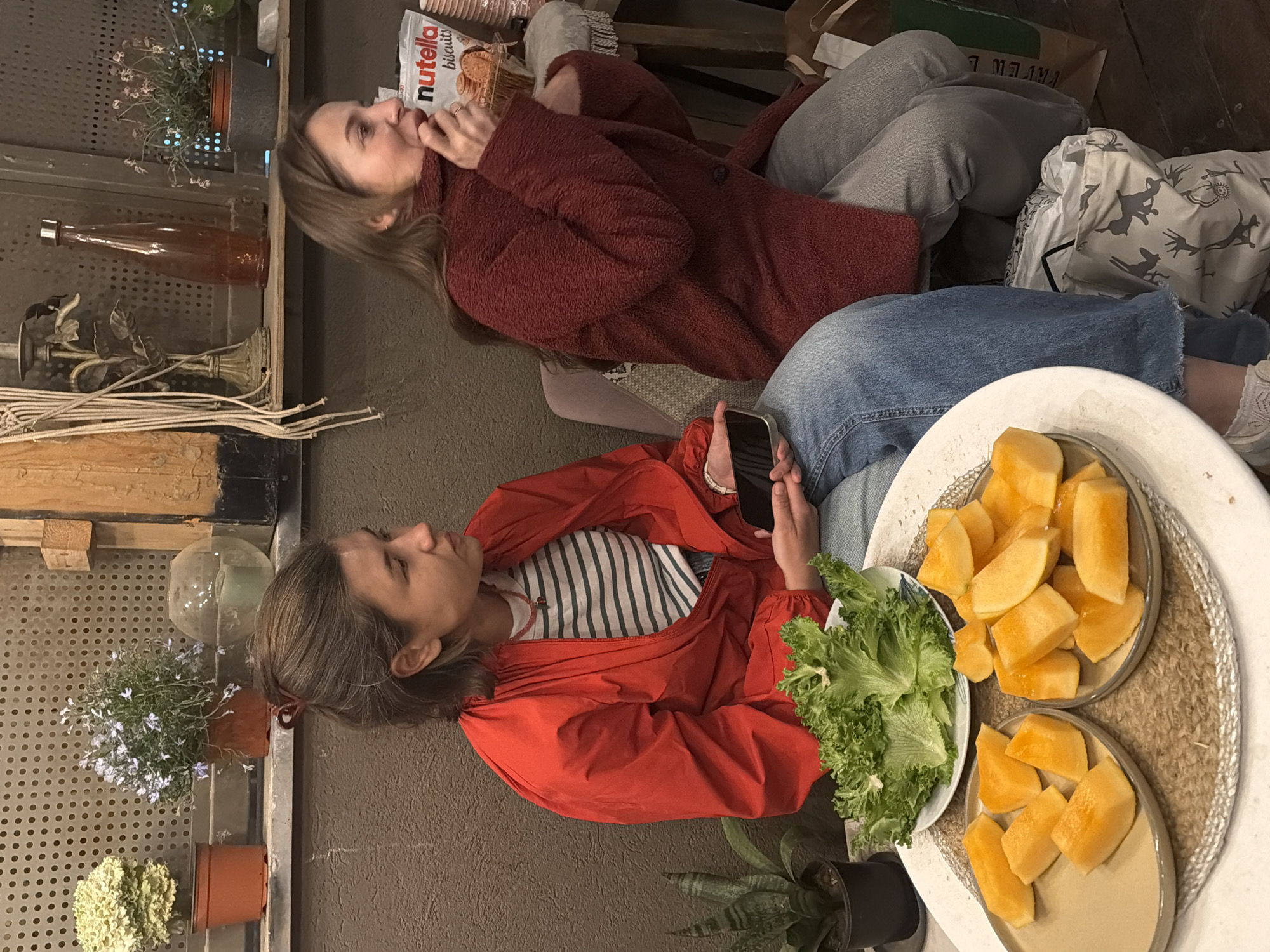 This media file captures two young women relaxing indoors, likely in a casual cafe or living space in Tel-Aviv, Israel.
The woman on the left is reclining on what appears to be a sofa, wearing a warm, red fuzzy jacket and light grey pants. She is looking upwards, and holding a bag of "Nutella biscuits" near her mouth, suggesting she is enjoying a snack. The text "Nutella biscuits" is clearly visible on the packaging.
To the right, a second woman is seated, wearing a bright red jacket over a striped shirt and blue jeans. She is holding a black smartphone and looking towards a low table in front of her.
The table holds a casual spread of food: two plates are generously filled with neatly cut pieces of cantaloupe, and a third plate contains fresh green lettuce. These are arranged on a textured, woven placemat.
The background features multi-tiered shelving with a perforated metal surface, adorned with several potted plants, some bearing small flowers, and a large, clear glass bottle. Another decorative item resembling a candle holder is also visible. The overall setting is cozy and informal, with soft, ambient lighting suggesting it is either daytime or early evening. Both women appear relaxed and at ease, enjoying their time together.