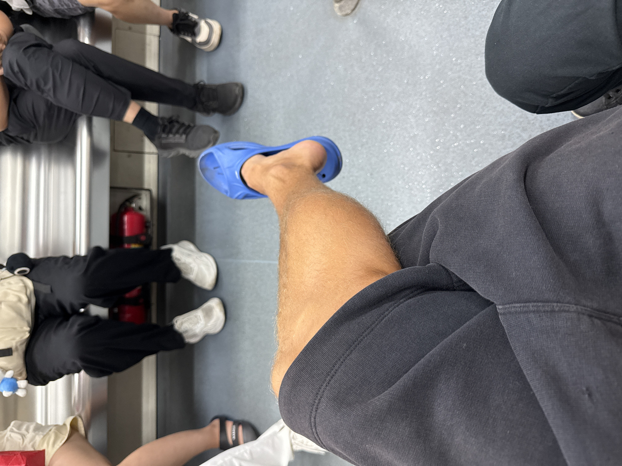 The image shows a close-up of a person's legs and feet, seated on a public transport vehicle that appears to be a train or subway. The person is wearing dark sweatpants and a blue Croc-style shoe on their right foot, with their left foot bare and resting on the shoe. In the background, other passengers are visible, also seated, with their legs and feet showing. One passenger on the left is wearing white sneakers, while another in the center wears black sneakers. The seats appear to be made of a metallic material, and the floor is a light-colored, textured surface. Red containers are visible under the seats in the background, possibly fire extinguishers. The overall environment suggests an indoor, public transit setting within the city of Haizhu District, China.