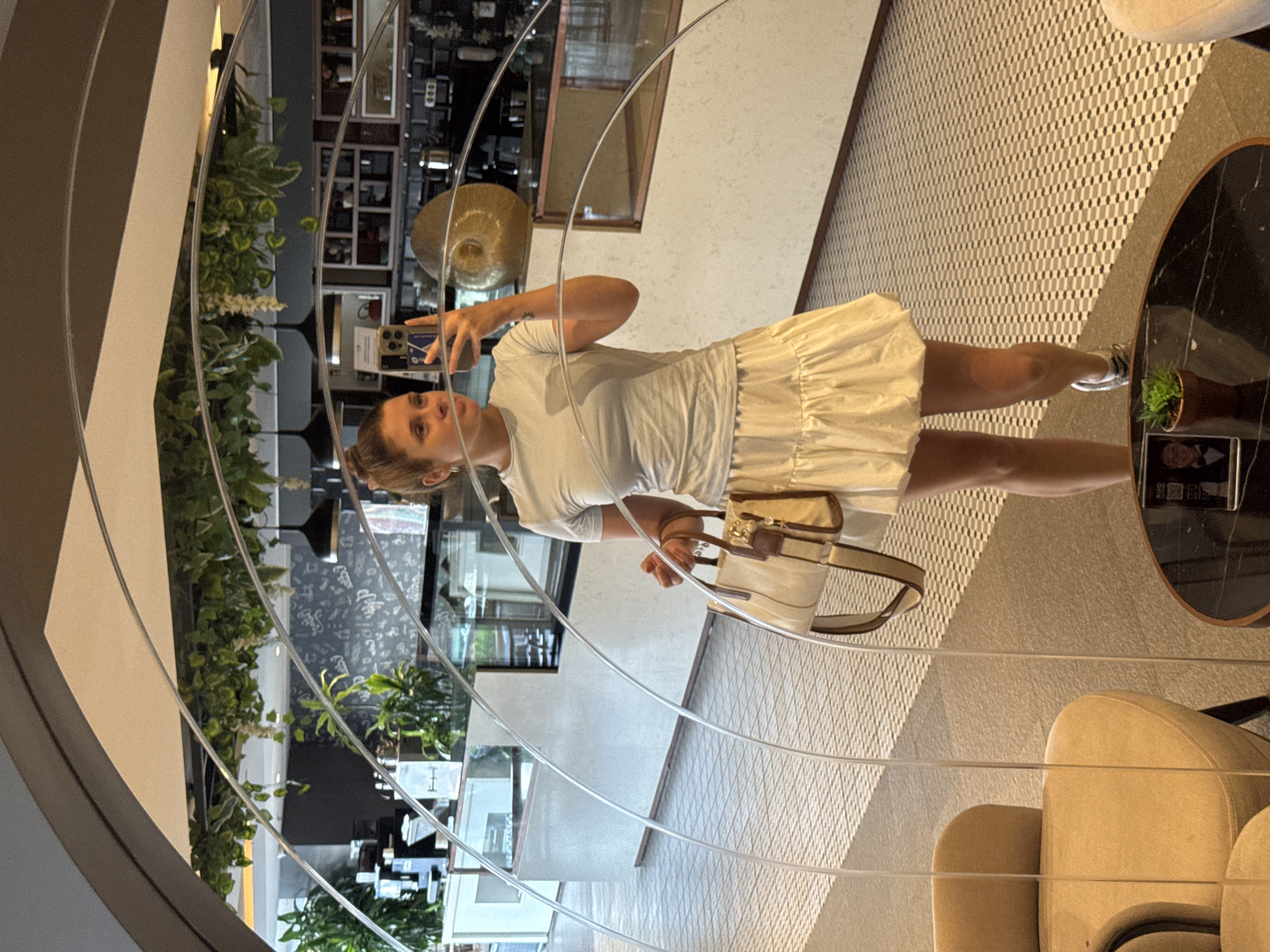 A woman is standing in what appears to be a cafe or restaurant, taking a mirror selfie. She is wearing a light blue t-shirt and a cream-colored tiered skirt. She is holding a cream and brown handbag in her left hand and her phone in her right hand. The background shows a counter with coffee machines and shelves filled with various items. The floor is covered in a black and white tiled pattern. The environment is modern and chic, with hanging plants and a large, curved mirror that distorts the reflection. The woman's hair is pulled up in a bun, and she is looking at her phone. The scene appears to be set in the city of Aksu, Turkey.