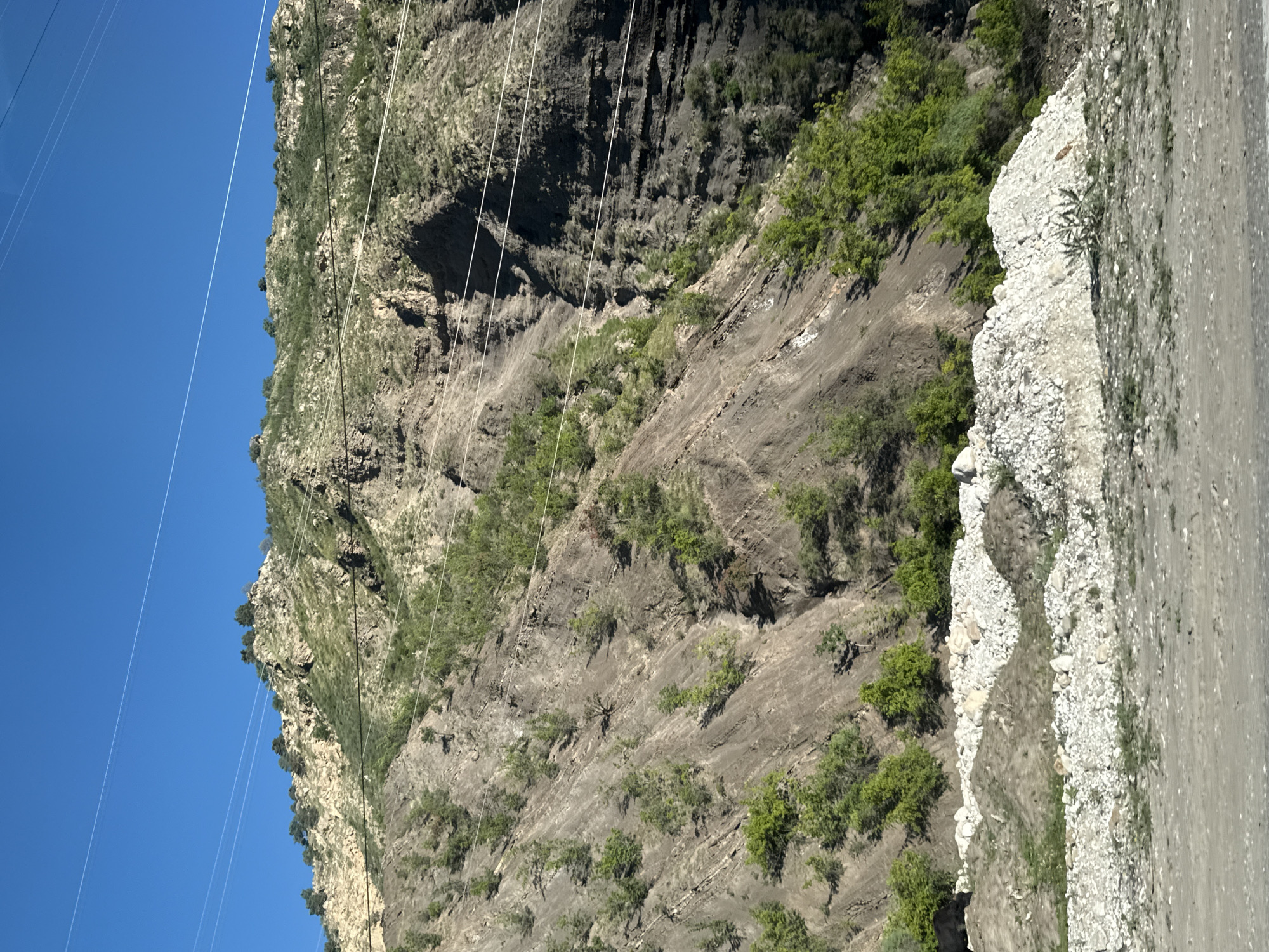 This media file captures a rugged mountainous landscape in Gimry, Russia, under a clear, bright blue sky. The dominant subject is a steep, rocky mountainside, characterized by dark grey and brown rock formations with visible stratifications and textures. Sparse green vegetation, including small bushes and patches of grass, is scattered across the slopes and crevices of the mountain.
Several thin, white power lines traverse the scene diagonally, stretching across the expanse of the sky and over the mountainside, suggesting human infrastructure in this natural environment.
To the right of the mountainside, an unpaved dirt road or path is visible. It runs roughly parallel to the slope. Between the road and the base of the mountain, there is a distinct bank composed of white, light-colored rocks and gravel, creating a visual contrast with the darker hues of the mountain and the road.
The image was taken during daylight hours, indicated by the vibrant blue sky and uniform illumination across the landscape, suggesting clear and sunny weather. There are no people, animals, or visible text in the image. The scene conveys a sense of a remote, natural setting, albeit one that is intersected by human utilities and pathways.