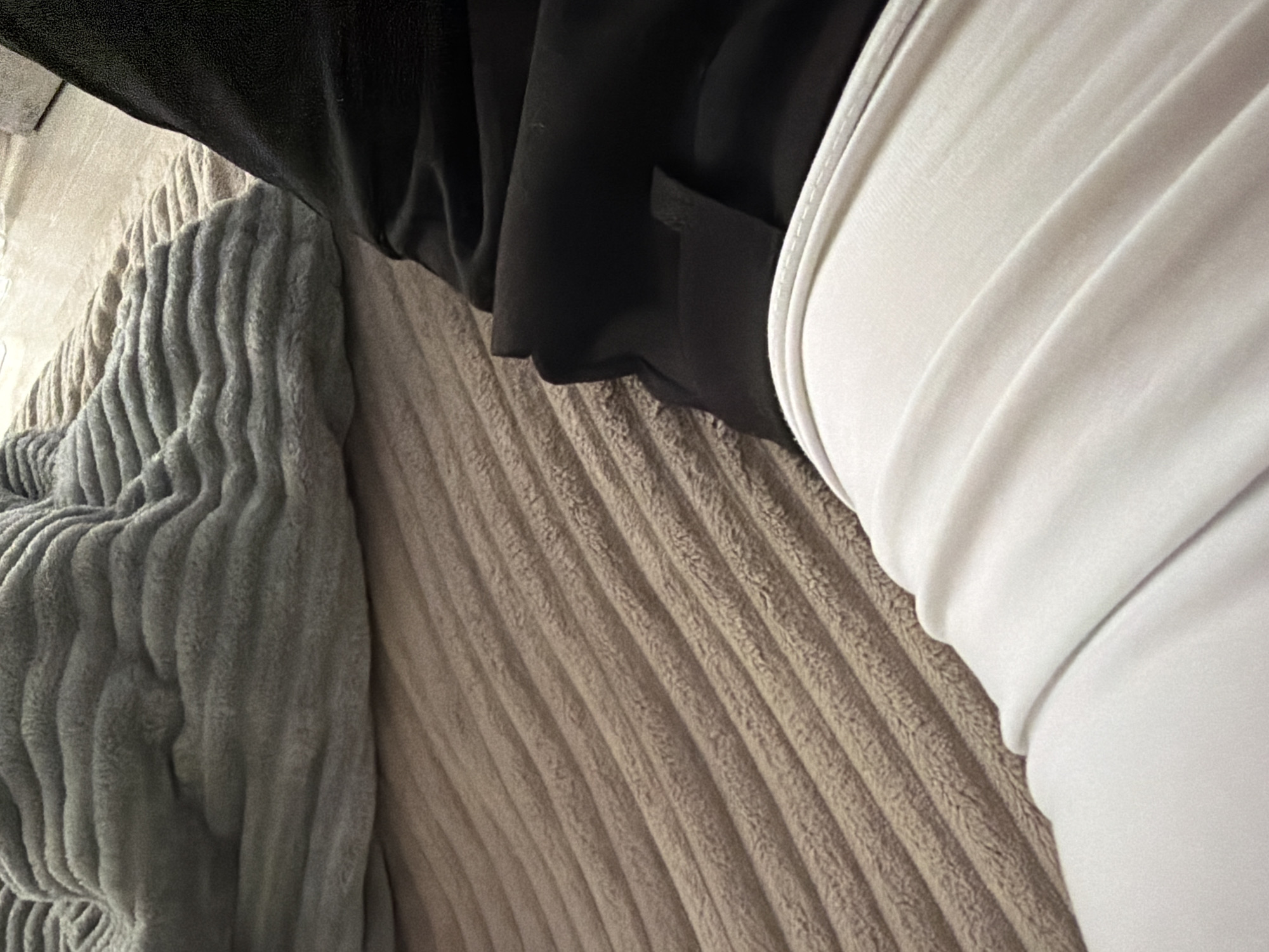 The image shows a close-up of a person sitting or lying down on a beige corduroy surface. They are wearing a black jacket or shirt with a pocket, and a white garment. A gray, textured blanket rests near the left side of the corduroy surface.