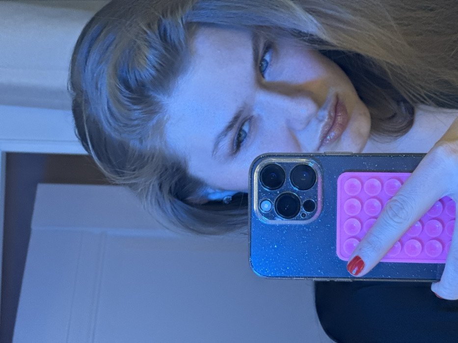 A person with light hair and blue eyes poses for a selfie in a mirror. They hold a dark, speckled smartphone with four camera lenses and a pink, circular textured grip on the back. The person's red-painted fingernail rests on the grip. The lighting appears blue, casting a cool tone on the image. A partially visible white doorframe is in the background.