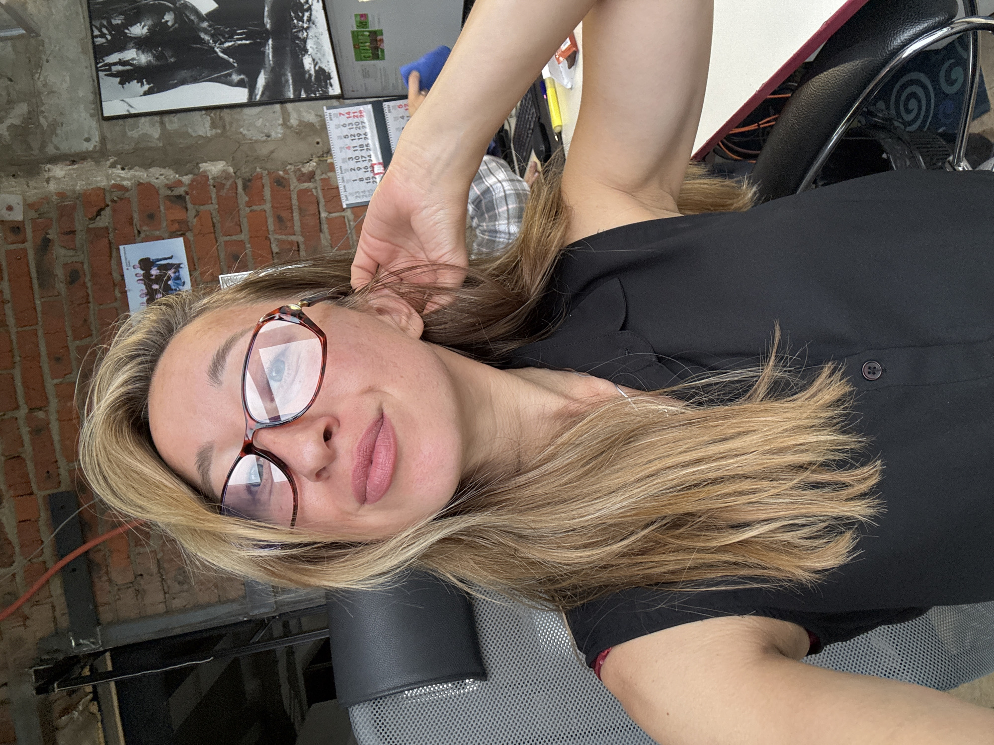 The image shows a close-up selfie of a woman with blonde hair, wearing glasses and a black sleeveless top. She is sitting in an office chair, with a brick wall and office equipment visible in the background. The scene appears to be an indoor office environment, possibly in the city of Chaikovsky, Russia, given the context provided. The woman is looking towards the camera, and her left arm is raised, touching her hair. A calendar is visible on the wall, showing dates for June and July. There is also a framed picture on the wall, and a computer monitor in the background.