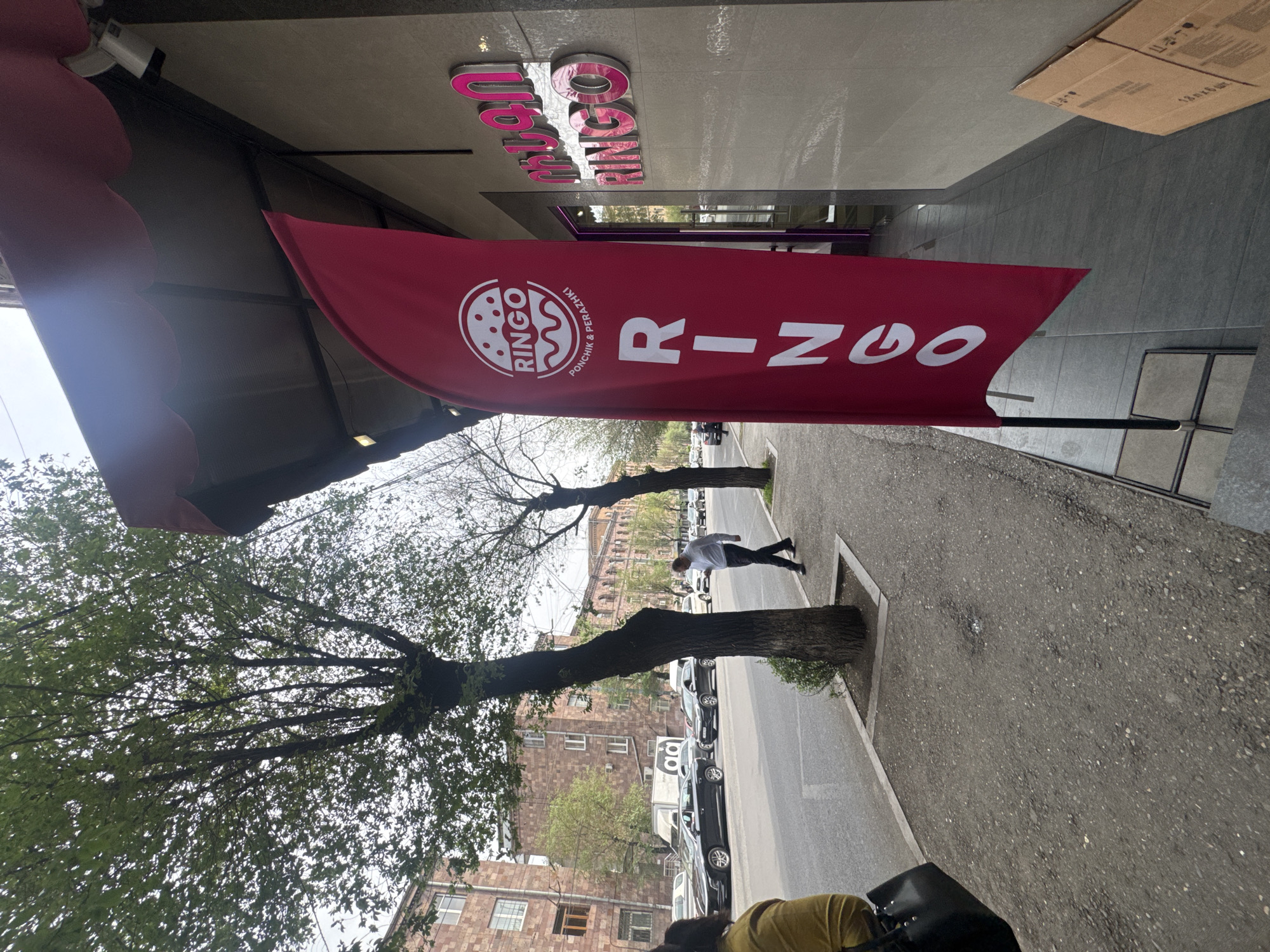 The image depicts a street scene in Yerevan, Armenia, with a "RINGO" bakery advertisement prominently displayed. The advertisement is a red banner with the word "RINGO" in white letters and a logo of a donut on the left side. Above the banner is a sign for "RINGO" in pink lettering.
The street is lined with trees and buildings. Cars are parked along the side of the road. A person is seen walking on the sidewalk. The image is taken from a slightly elevated perspective.