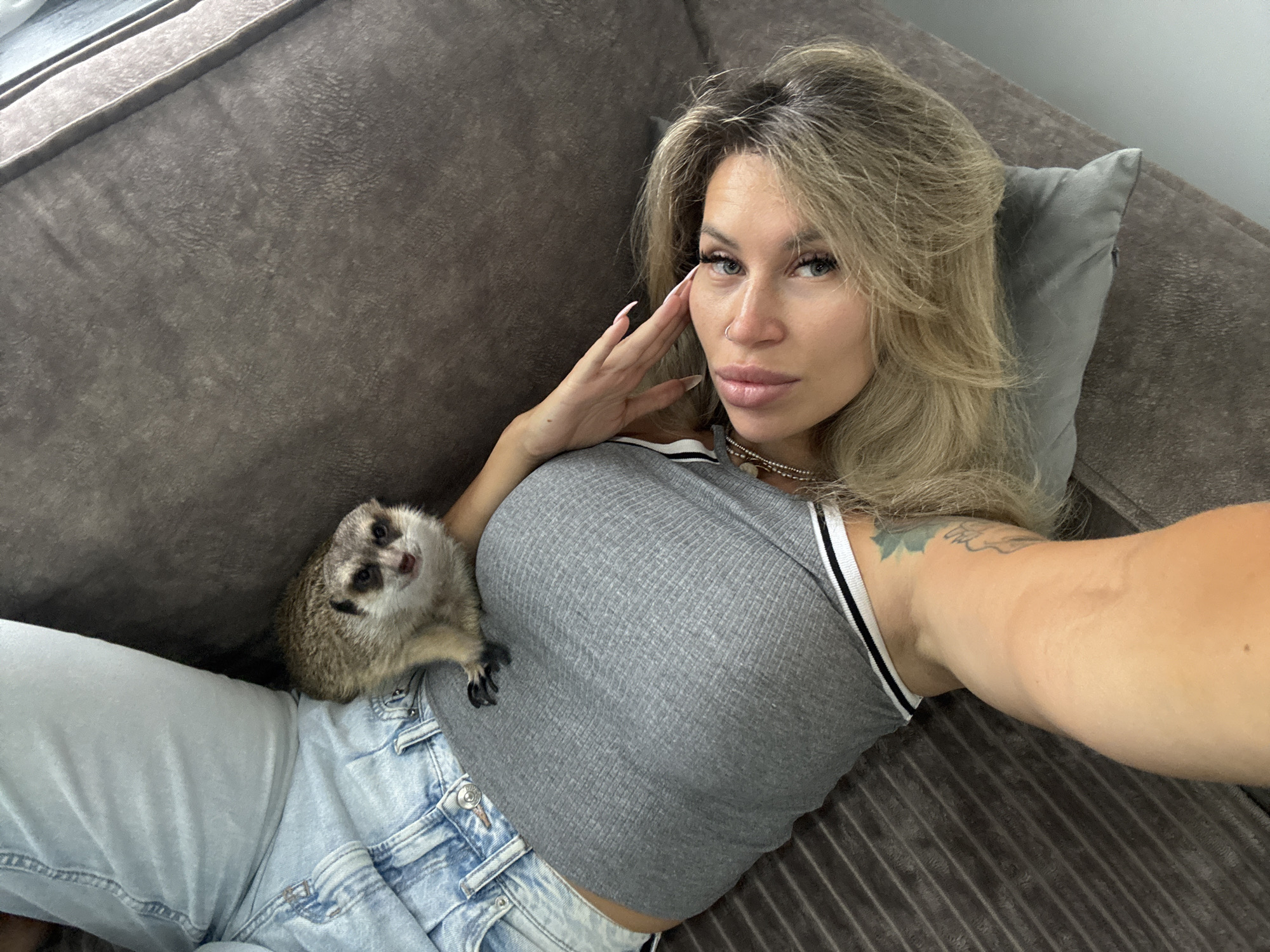 An eye-level selfie shows a blonde woman and a meerkat resting on a dark grey, textured sofa indoors, likely in a living room in Moscow, Russia. The woman is positioned on her back, her head resting on a light grey pillow. She has light eyes, long manicured nails, a nose piercing, and a tattoo on her left upper arm. She wears a grey ribbed crop top with a white and black striped trim and light blue denim jeans. Her expression is posed, looking directly at the camera, with one hand gently touching her face. A small, furry meerkat with distinctive black markings around its eyes is perched on her chest and lap, also looking towards the camera with an alert expression. The overall lighting suggests it is daytime. No text is visible in the image.