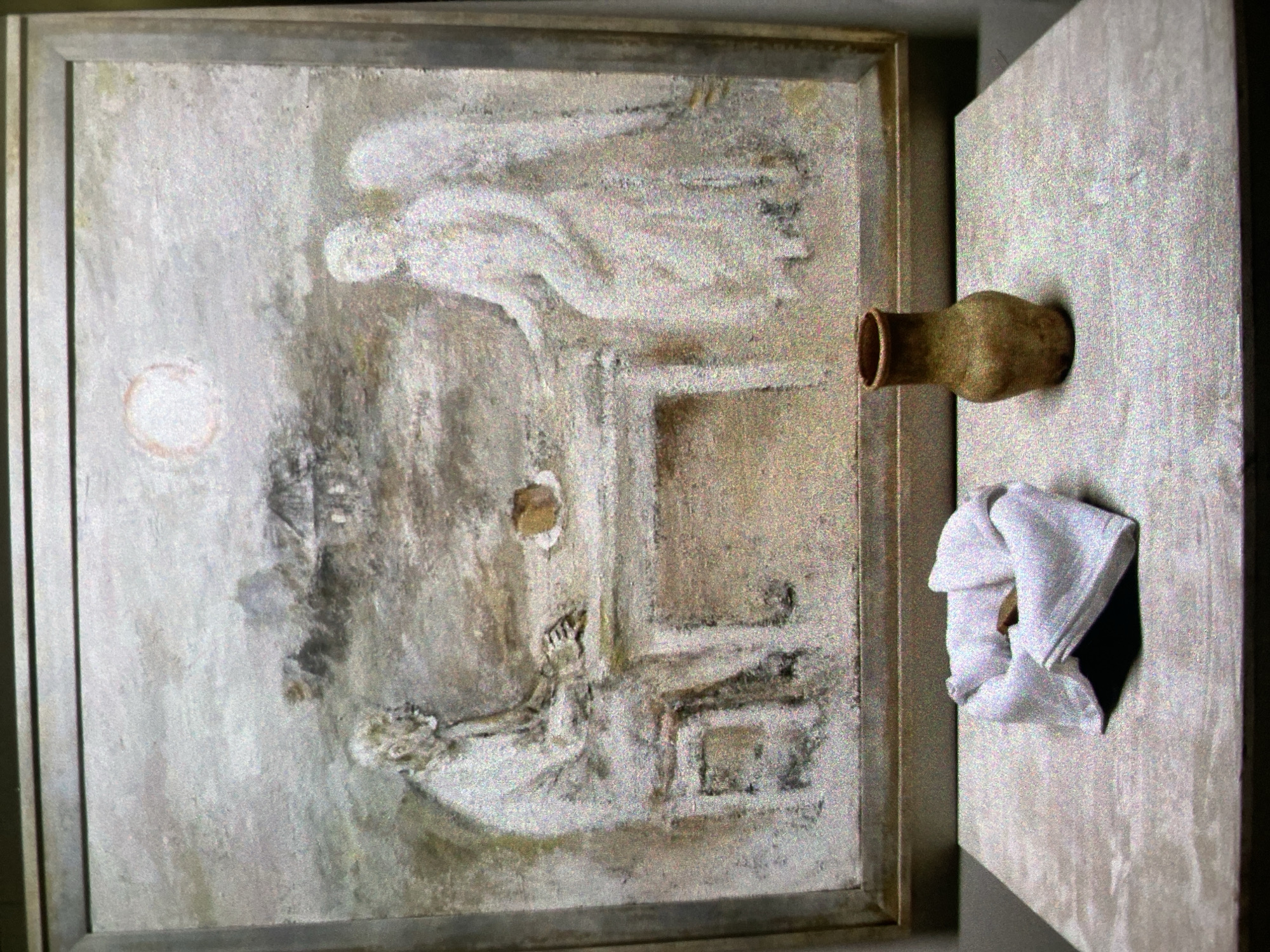 The image features a framed painting depicting a scene with two figures, possibly an angel tending to a seated or reclining figure, beneath a circular, sun-like object. The painting is executed in a muted, monochromatic palette. To the right of the painting, a pottery vessel is affixed to a vertical surface. Below the vessel, a folded white cloth is stuffed into a small, dark cavity.
The location in the city of Saint Petersburg, Russia is not discernible from the image alone.