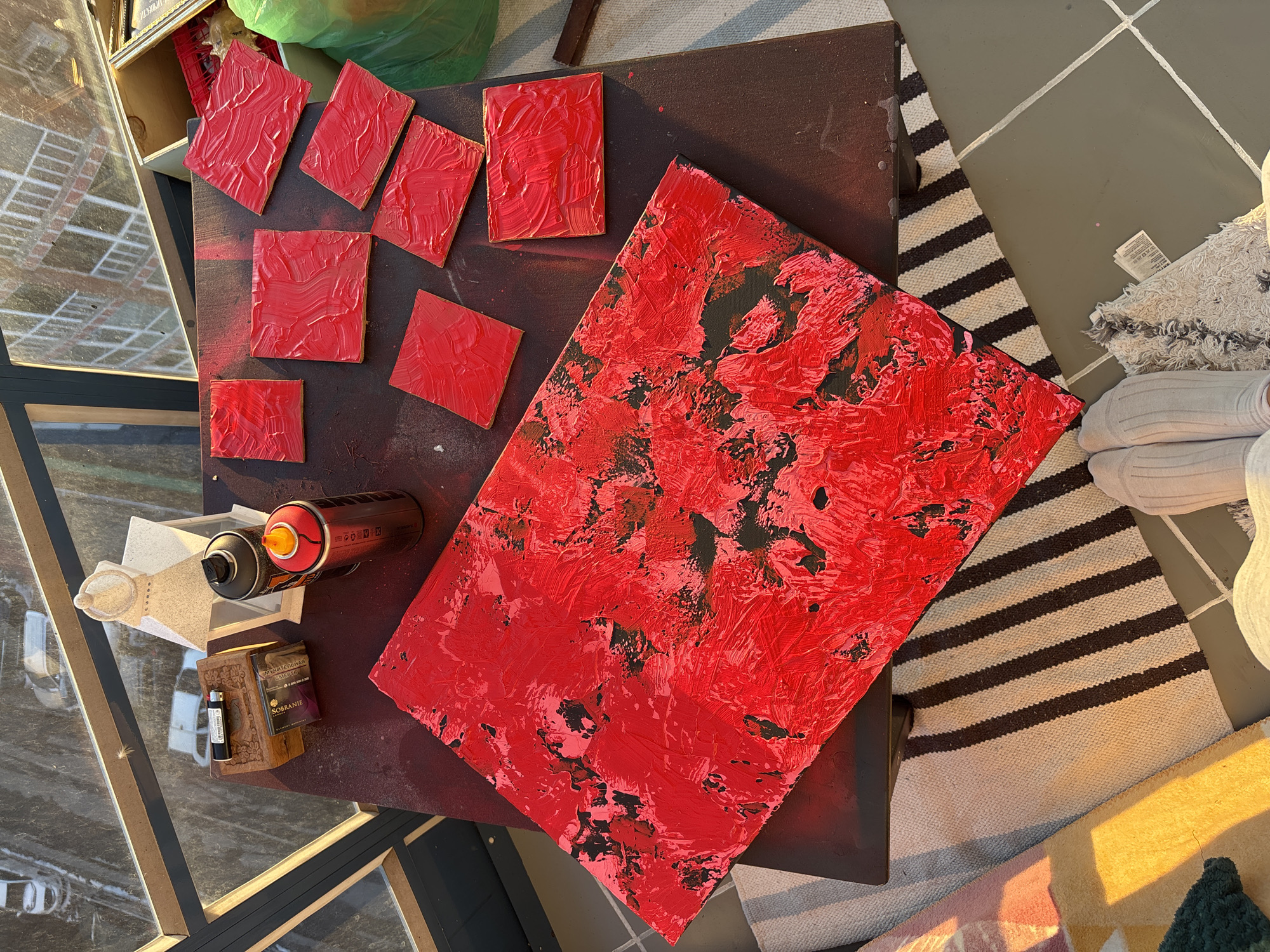 An overhead shot displays a maroon-colored table or surface covered with art supplies and art pieces, alongside a glimpse of what seems to be an interior setting and potential city view through a window.
On the table, there are several small, square-shaped canvases covered in thick red paint, positioned alongside a larger rectangular canvas, also covered in a thick layer of red paint, with some black paint showing through. A pair of spray paint cans, one black and one red, along with a wooden box containing cigarettes and a lighter, sit near the canvases.
In the background, there's a portion of a striped rug and a person's feet in grey socks are visible. Through a window, a city view is suggested by the grid-like pattern, reflecting what looks like buildings.
Based on cigarette brand and the language on the cigarette box, the city could be Krasnoyarsk, Russia.