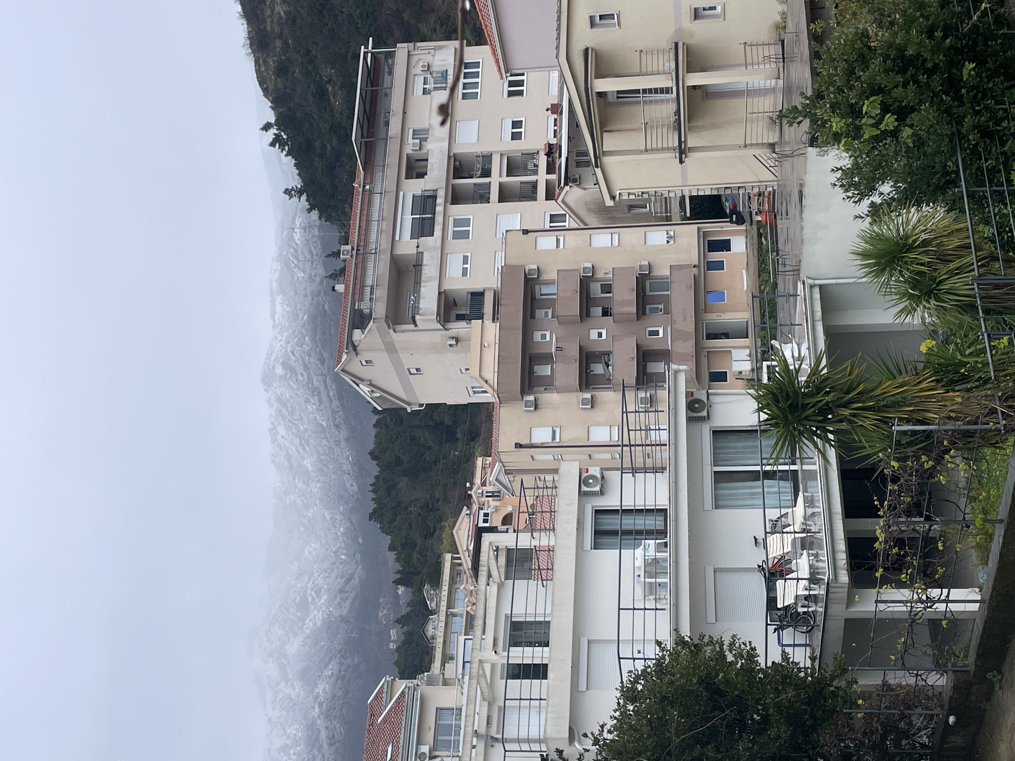 The image depicts a residential hillside area in Budva, Montenegro, under an overcast sky. In the background, snow-capped mountains are partially obscured by low-hanging clouds or mist, with their lower slopes densely forested.
The scene is dominated by multiple modern residential buildings of varying heights and architectural styles, constructed on a sloped terrain. Buildings feature light-colored facades (white, beige, light brown, pinkish-beige), red-tiled or flat roofs, and numerous balconies with dark metal railings.
In the immediate foreground, a dark metal fence is partially visible, with green foliage including small shrubs and a palm plant. A prominent white multi-story building on the left has a large balcony equipped with two white lounge chairs and a red bicycle. This balcony is fitted with glass sliding doors and white roller shutters. An external air conditioning unit is affixed to its facade.
Moving into the mid-ground, a cluster of residential buildings is visible. A multi-story beige building on the right features several balconies with similar railings. Another taller beige building towards the center has multiple balconies, some enclosed, and numerous windows. Air conditioning units are prevalent on the exterior walls of most buildings. One building displays rooftop solar panels. A dark-colored vehicle is partially visible between two buildings further back on the right.
No people or discernible actions are present in the image. The environment suggests a temperate climate with recent snowfall at higher elevations.