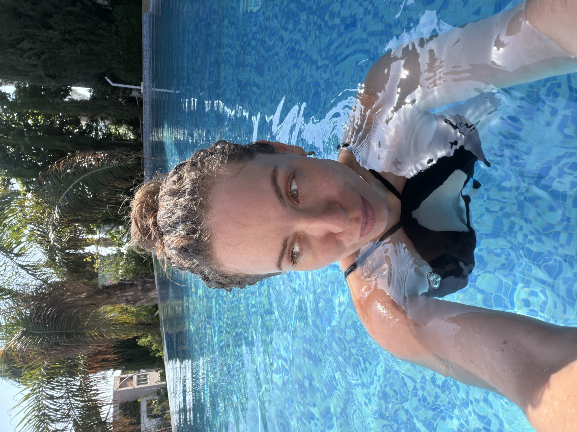 A woman is in a swimming pool. She is looking at the camera, with her hair pulled up. She is wearing a black and white bikini top, with a sheer white garment over it. The water is a bright blue, with sunlight reflecting off the surface. Green trees and a building are visible in the background, suggesting an outdoor location. The scene appears to be set during the day, under sunny weather. The woman's expression is relaxed and content.