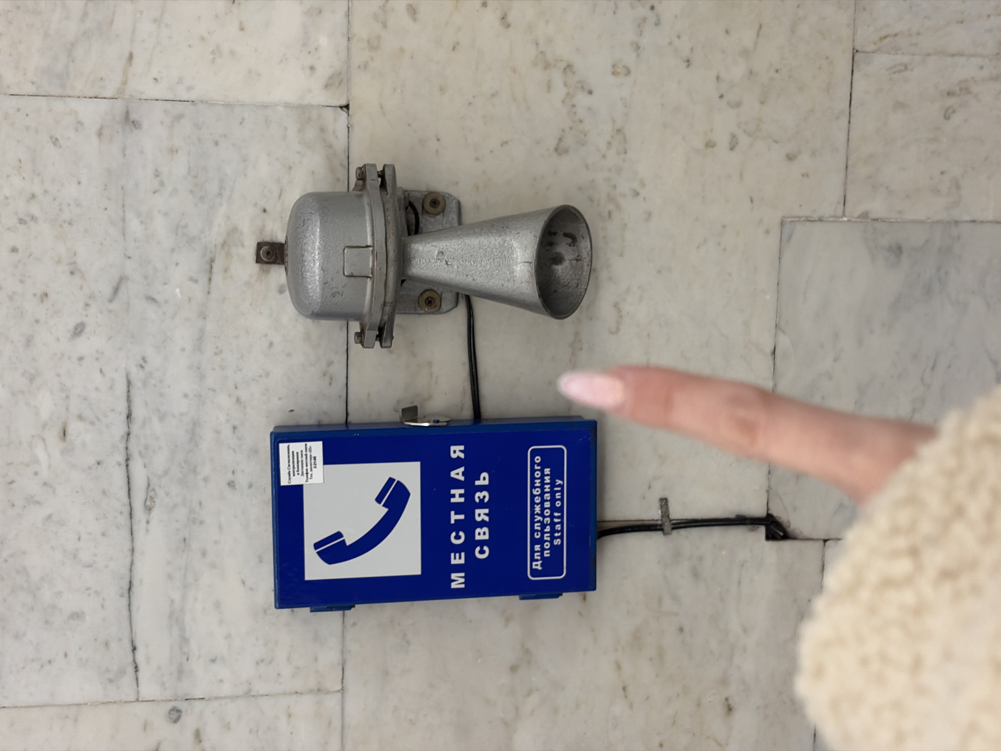 A wall-mounted phone, identified by the inscription "МЕСТНАЯ СВЯЗЬ" (Local Communication) and "Для служебного пользования. Staff only," is affixed to a marble wall. A gray horn speaker is mounted above the phone. A finger is pointing towards the phone. The location is Saint Petersburg, Russia.