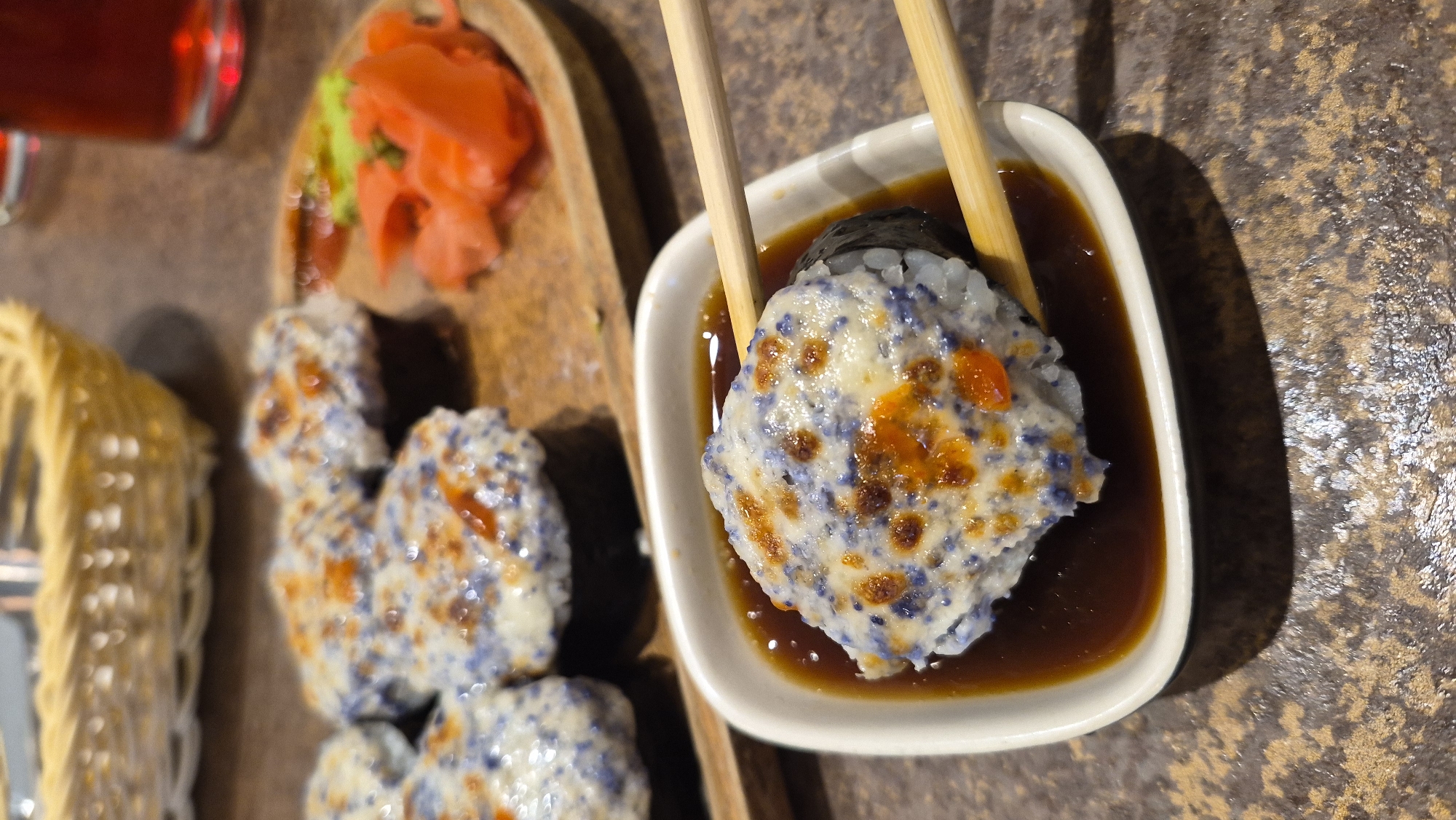 A close-up shot captures a piece of sushi being dipped into soy sauce with chopsticks. The sushi roll features rice, a dark seaweed wrap, and a generous topping of a creamy white sauce speckled with blue and orange elements. In the background, several other pieces of the same sushi are arranged on a wooden platter alongside pickled ginger and wasabi. A woven basket and a glass of red beverage are also visible in the background. The image appears to be taken in a restaurant setting, possibly in Daugavpils, Latvia, given the context of the request.
