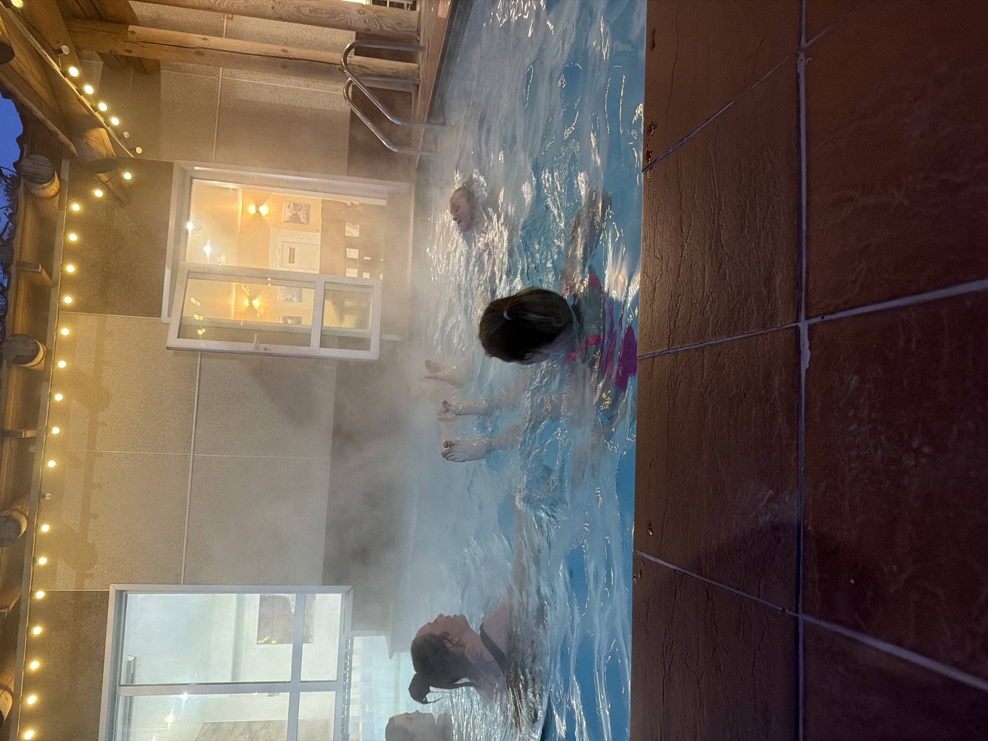 The image shows a group of people in an indoor pool with steam rising from the water. The pool is surrounded by tiled walls. There are windows on one side of the pool and string lights above. People are relaxing or swimming in the water. The location appears to be the Demidkovo SPA Hotel in the city of Chaikovsky, Russia.