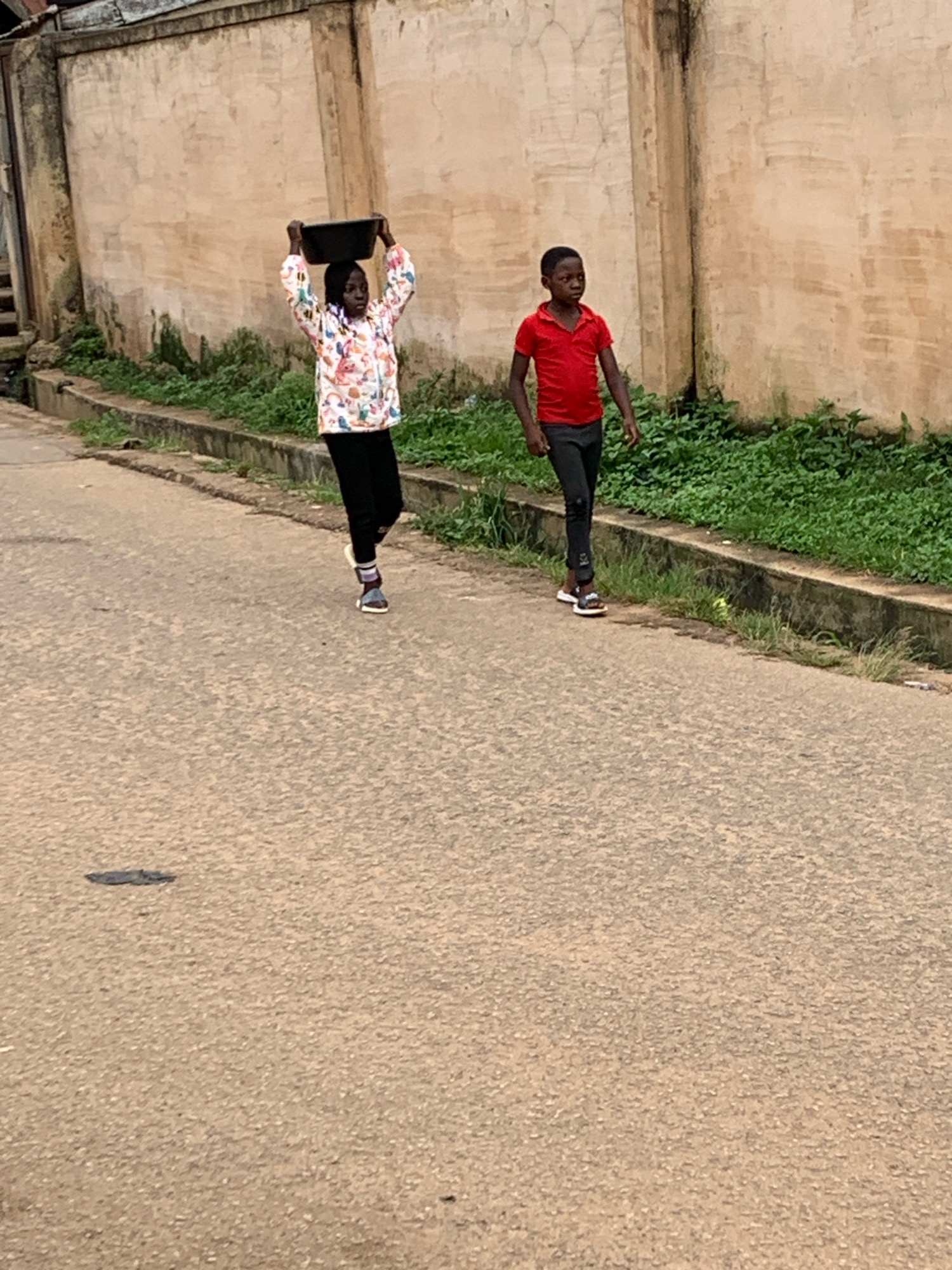 Two children are walking on a paved road in Jos, Nigeria. On the left, a young girl wearing a colorful jacket and black pants carries a dark basin on her head. On the right, a boy in a red shirt and black pants walks alongside her. A textured, light-colored wall with some weathering and greenery running along its base forms the background. The road surface appears to be asphalt.