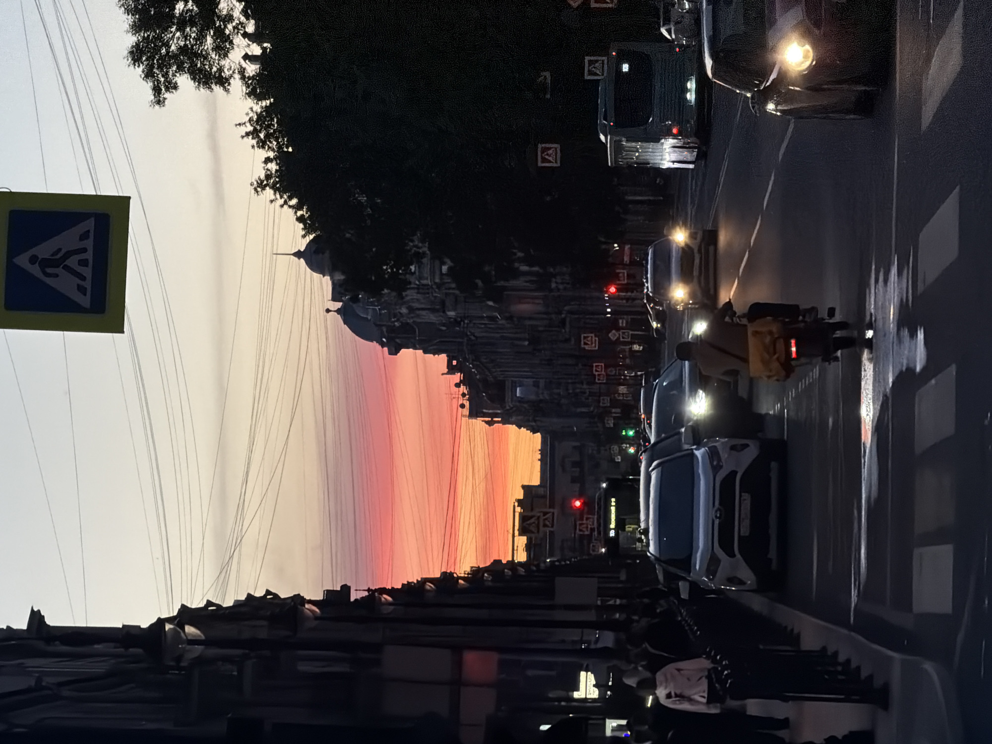 The image depicts a street scene in Saint Petersburg, Russia, during a sunset. The sky displays vibrant hues of orange and pink. On the left side of the street, dark buildings with ornate architectural details line the road, interspersed with streetlights. A pedestrian walkway with a crosswalk is visible in the foreground. Several cars are on the road, including a white SUV in the center, a bus with illuminated lights on the right, and a motorcycle with a yellow bag on the back. People are walking on the sidewalk on the left. A pedestrian crossing sign is visible hanging over the street. The overall atmosphere suggests a busy urban environment transitioning into evening.
