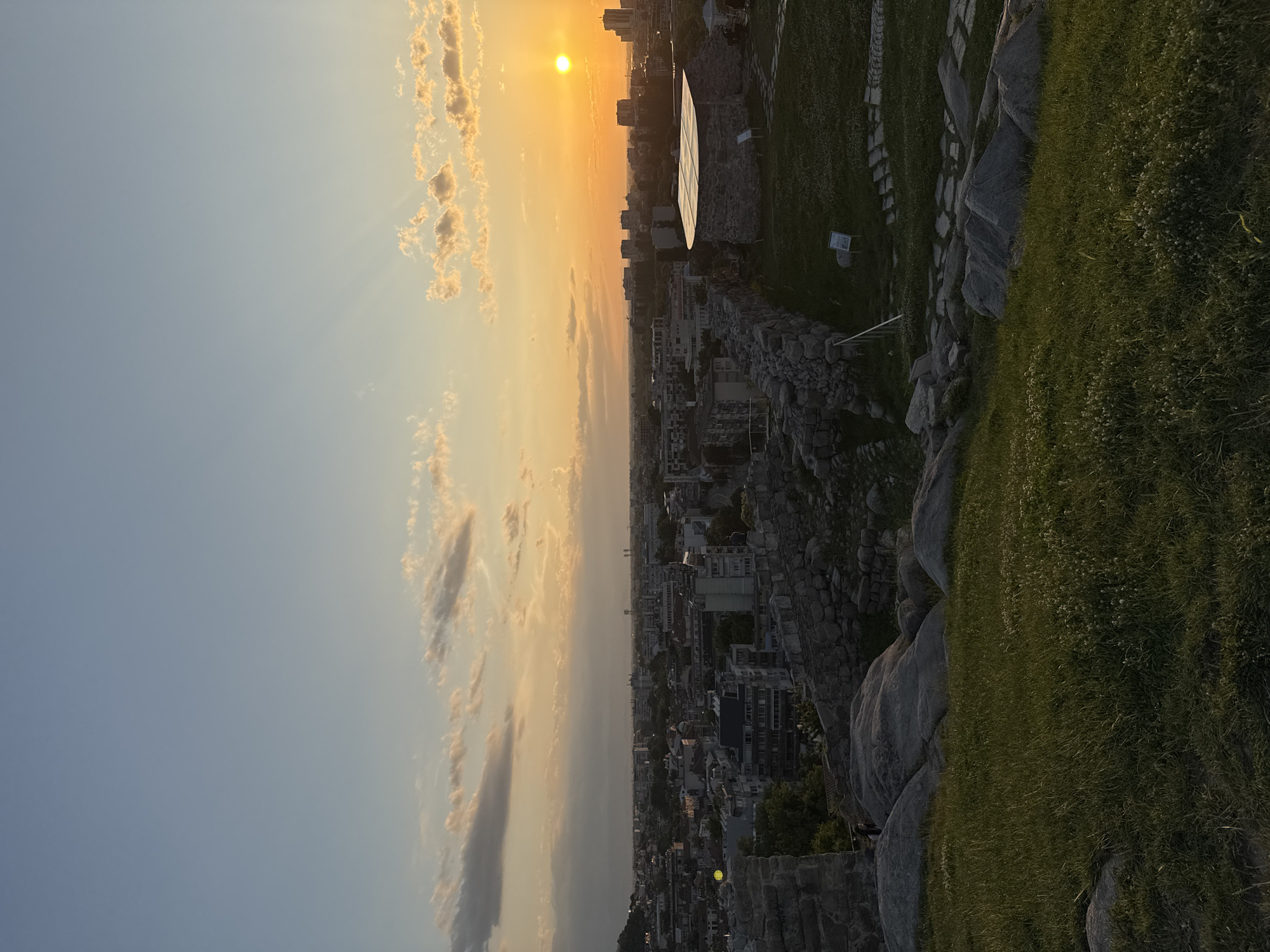 The image presents a panoramic elevated view of the city of Plovdiv, Bulgaria, at sunset. The sky displays a gradient from light blue at the zenith to an intense golden orange at the horizon, where the sun, a distinct bright orb, is positioned in the upper right. Crepuscular rays are visible emanating from the sun, piercing through scattered clouds that range in color from white to grey.
In the immediate foreground, a grassy slope is visible, featuring several large, smooth grey boulders. To the left and right, segments of ancient stone walls, constructed from irregular grey blocks, mark the edge of an elevated historical site. On the right, a small, dark stone building with a lighter, possibly modern, domed roof is situated on a patch of grass. A winding stone path, made of flagstones, leads around this structure, and small informational signs are present nearby.
The midground reveals the sprawling urban landscape of Plovdiv, densely packed with multi-story buildings of various architectural styles, including residential and commercial structures. Green spaces and tree lines are interspersed throughout the city. The cityscape extends towards the background, where a hazy horizon line is visible, with faint outlines of distant hills or mountains. Minimal human presence is indicated by very small, indistinct figures near the ancient walls on the left and potentially near the small building on the right.