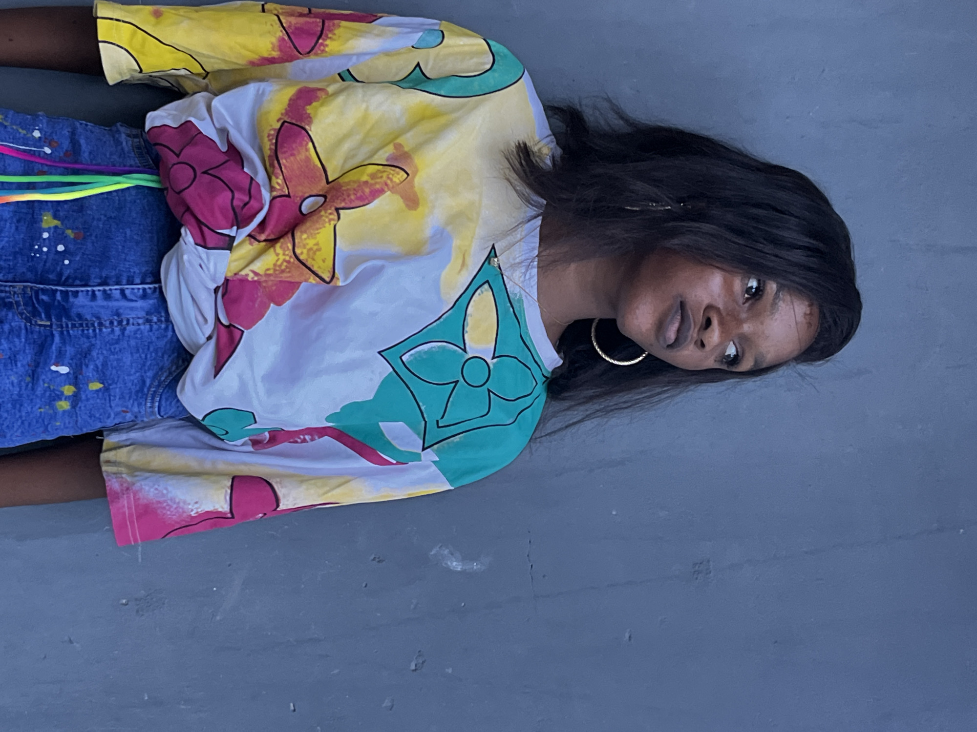 A woman with dark hair and brown skin wears a colorful, patterned shirt and denim shorts. She is positioned on a gray, flat surface. The image appears to be taken outdoors. The location in Badagry, Nigeria is not apparent from the image.