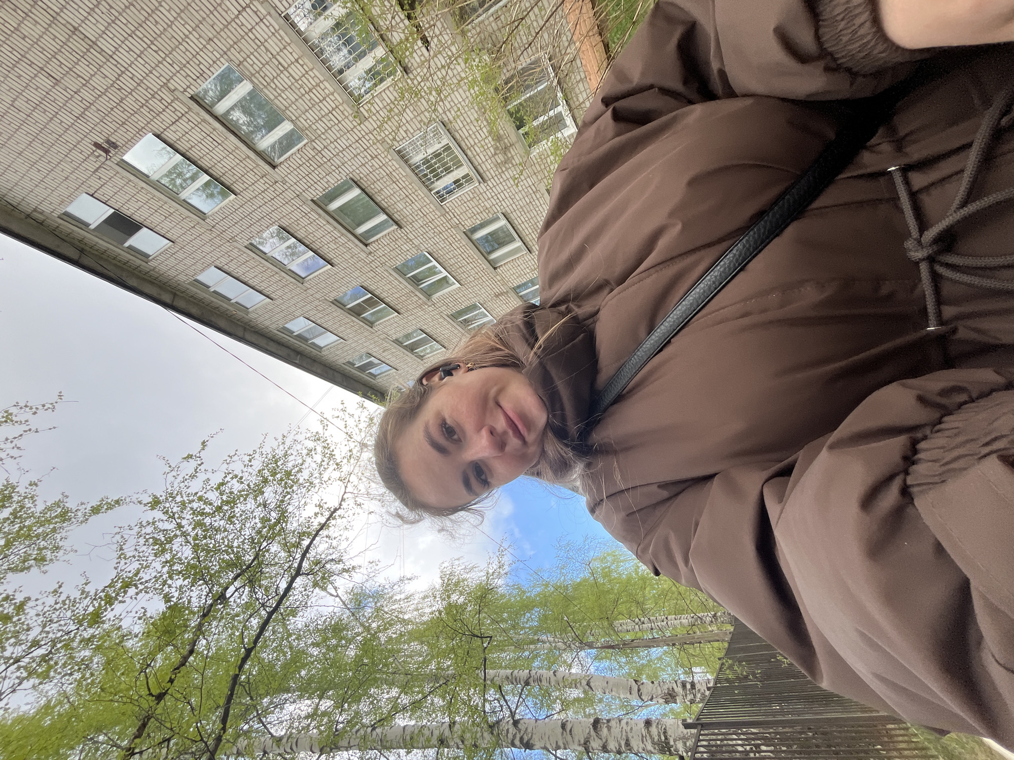 A woman with fair skin and light brown hair stands outdoors, looking down at the camera. She is wearing a brown puffer jacket with a black strap across her shoulder. Behind her, a tall, multi-story brick building with numerous windows rises into the frame. The building's façade is a light beige color. Below the building, tree branches with emerging green leaves reach upwards, and patches of blue sky are visible between the branches. The picture's perspective gives the impression of looking upwards towards the woman and the building. Given the architectural style and the presence of birch trees, it is plausible that the location is Chaikovsky, Russia.