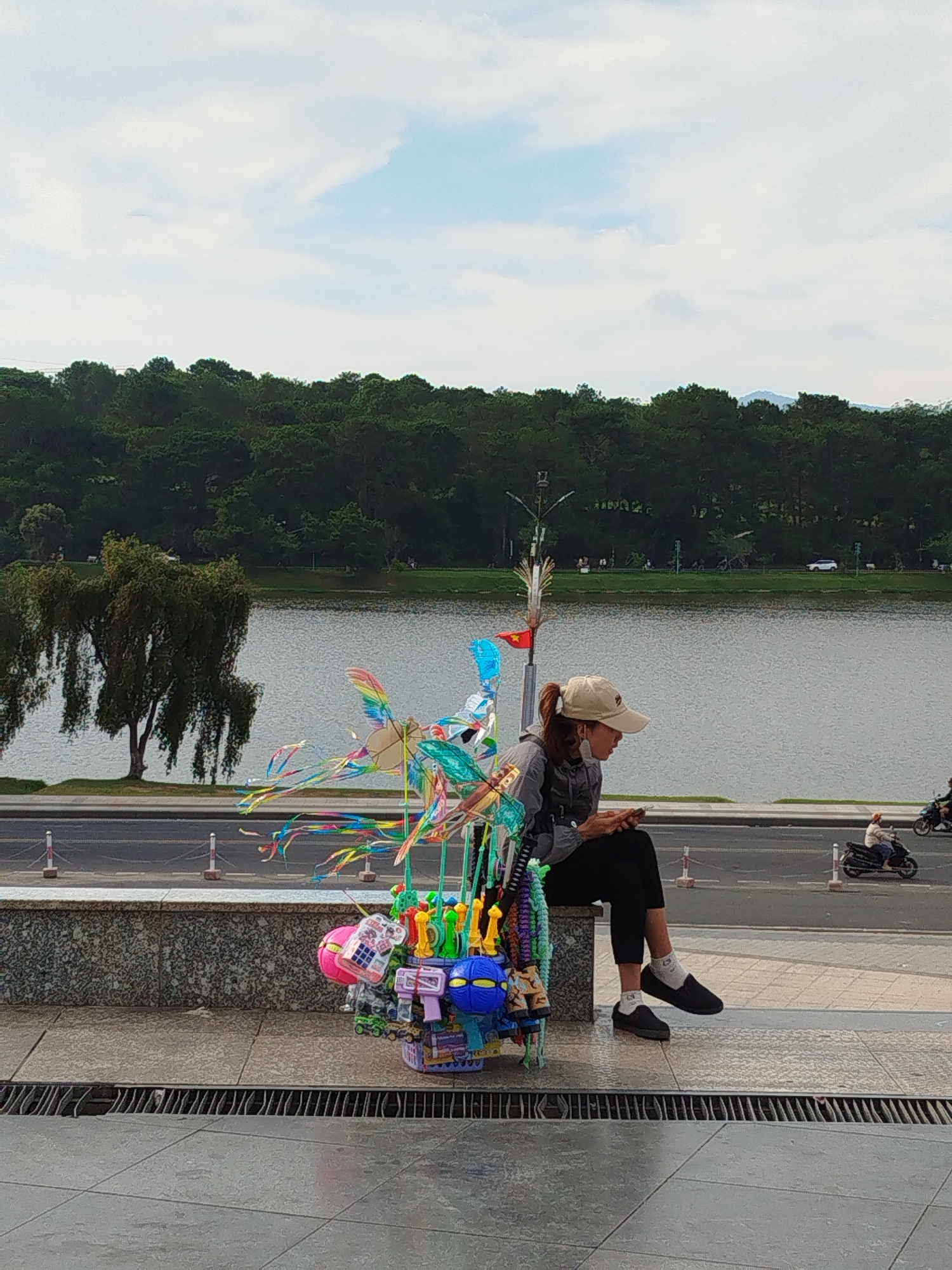 A medium shot shows a woman wearing a cap and mask sitting on a ledge with a collection of colorful toys next to her. She is using a smartphone. Behind her is a lake bordered by a road, with a forested area in the background. The sky is overcast with patches of blue.
Location: Da Lat, Vietnam, near Xuan Huong Lake.