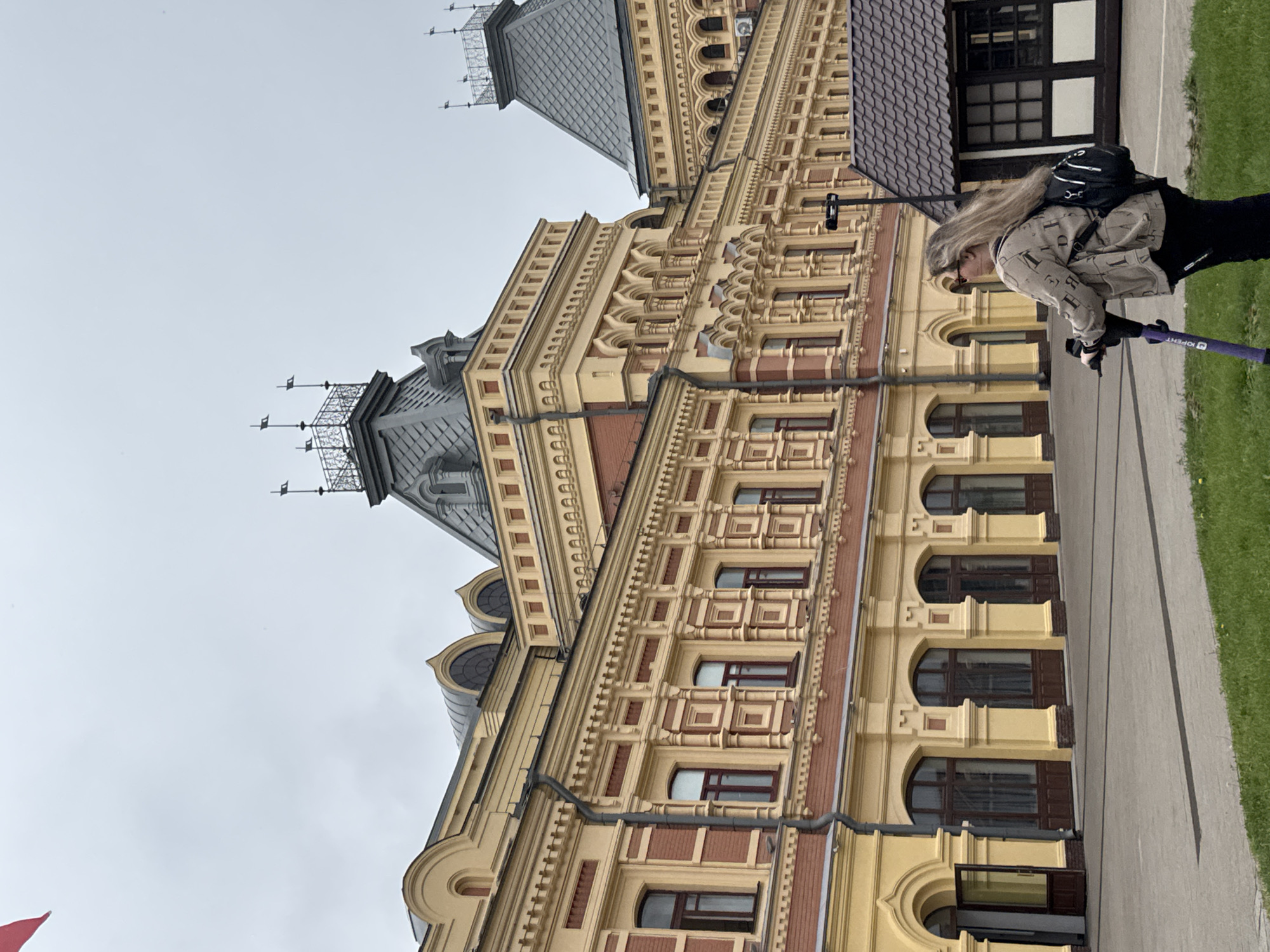 The image shows a woman with long blonde hair, wearing sunglasses and a patterned jacket, riding an electric scooter. She is on a paved area in front of a large, ornate building with yellow and red brick accents. The building has arched windows and decorative architectural elements. To the right of the building, there is a small, dark-roofed structure. The sky is overcast. This is likely the Nizhny Novgorod Fair (Nizhegorodskaya Yarmarka) in Nizhny Novgorod, Russia, due to the distinctive architecture of the buildings, which are characteristic of the fairgrounds.