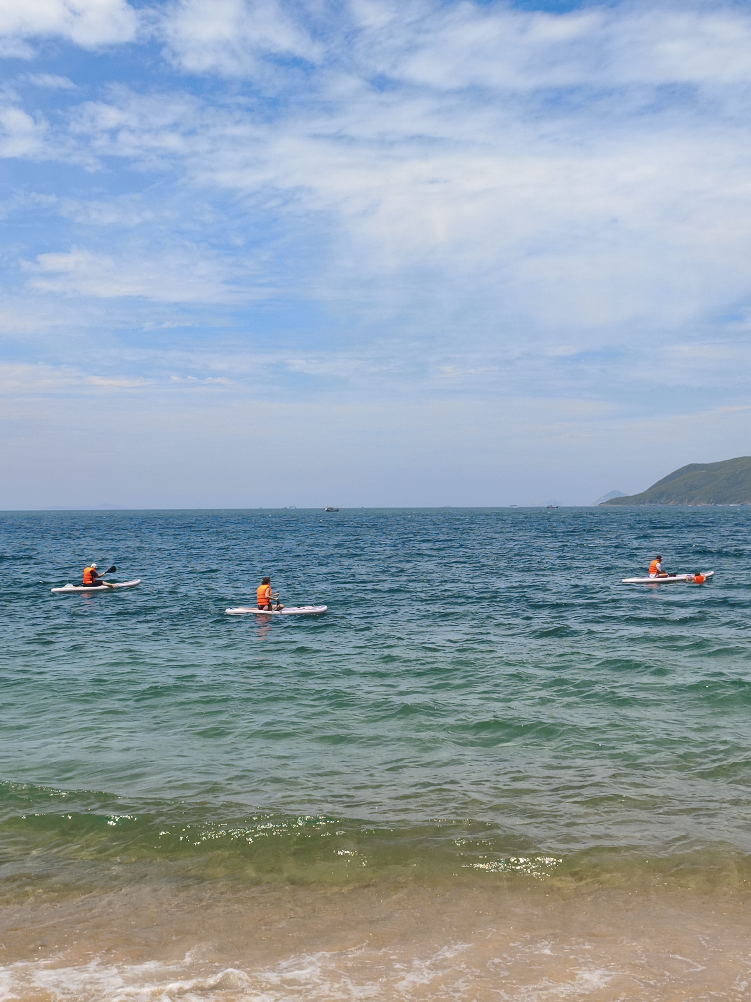 The image captures a serene view of the sea off the coast of Nha Trang, Vietnam, on a bright, partly cloudy day. In the foreground, gentle waves lap onto a sandy beach. The middle ground is dominated by the deep blue sea, with ripples indicating a light breeze. Three individuals are seen paddleboarding at varying distances from the shore. The closest paddleboarder is positioned towards the left side of the frame, facing left, while another is in the center, facing right. The third paddleboarder is further to the right, also facing right. All three individuals are wearing orange life vests, suggesting a recreational activity. In the background, the horizon is marked by a hazy sky with scattered white clouds and a distant, green, mountainous coastline on the right. Several small boats are visible on the water in the distance, adding to the sense of a coastal locality. The water transitions from a clear, shallow turquoise near the shore to a deeper blue further out.