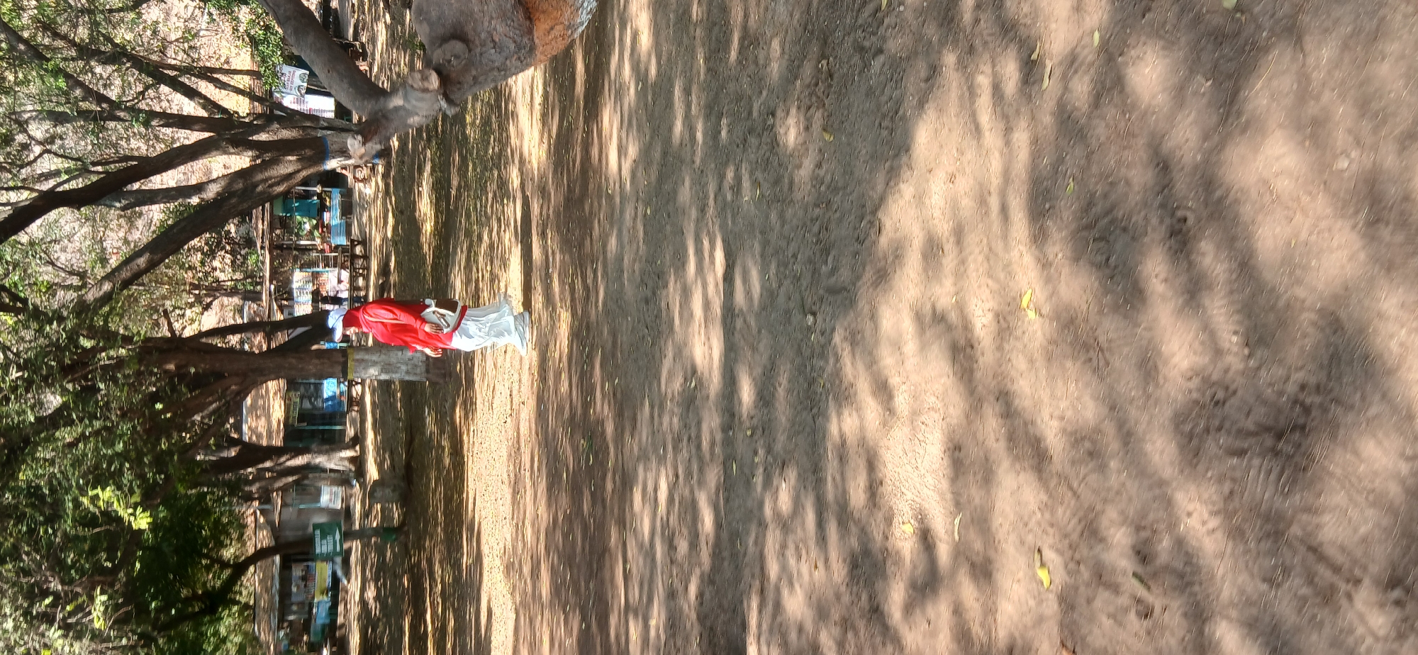 A person wearing a red top, white pants, and a white cap stands in a shaded area of a park. The person is looking to the left and has a bag over their shoulder. In the background, there are several small buildings with awnings and some tables and chairs. The ground is covered in dirt and fallen leaves, with shadows cast by trees. The location appears to be a public area, possibly a recreational park or rest stop, in Banyuglugur, Indonesia.