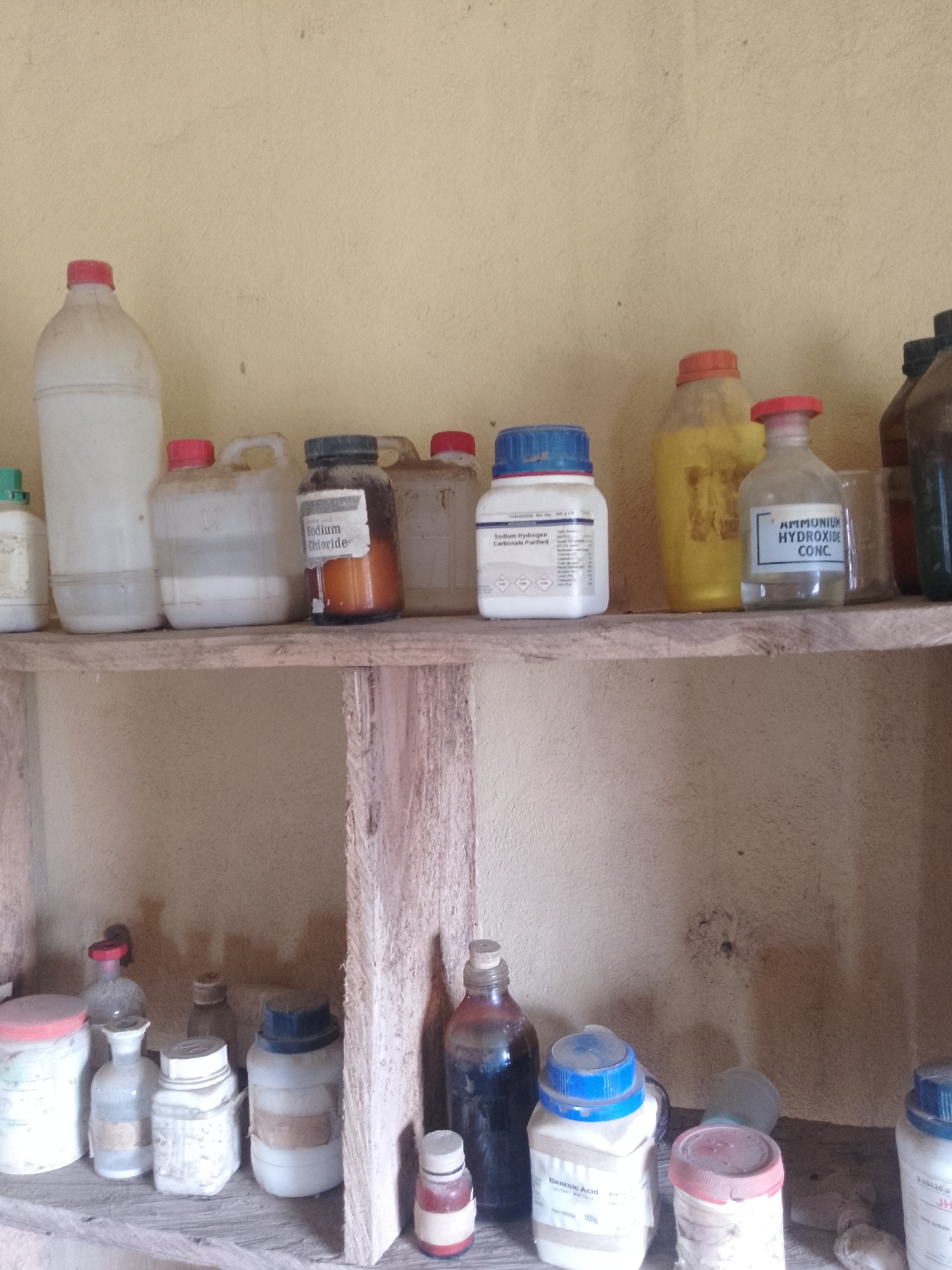The image depicts a dusty wooden shelf in what appears to be a storage area in Serti, Nigeria. The shelf is filled with various bottles and containers of chemicals, some of which are labeled. On the top shelf, from left to right, there are several large plastic bottles, a brown glass bottle labeled "Sodium Chloride", a yellow jug, a clear bottle labeled "Ammonium Hydroxide CONC.", and some other indistinct containers. The lower shelf holds a collection of smaller bottles and containers, many of which appear to be laboratory-grade chemicals. Some are labeled with handwritten tags, while others have printed labels that are difficult to read due to dust and age. The background wall is a light, textured color. The overall impression is one of an unkempt but functional storage space for chemical supplies.
