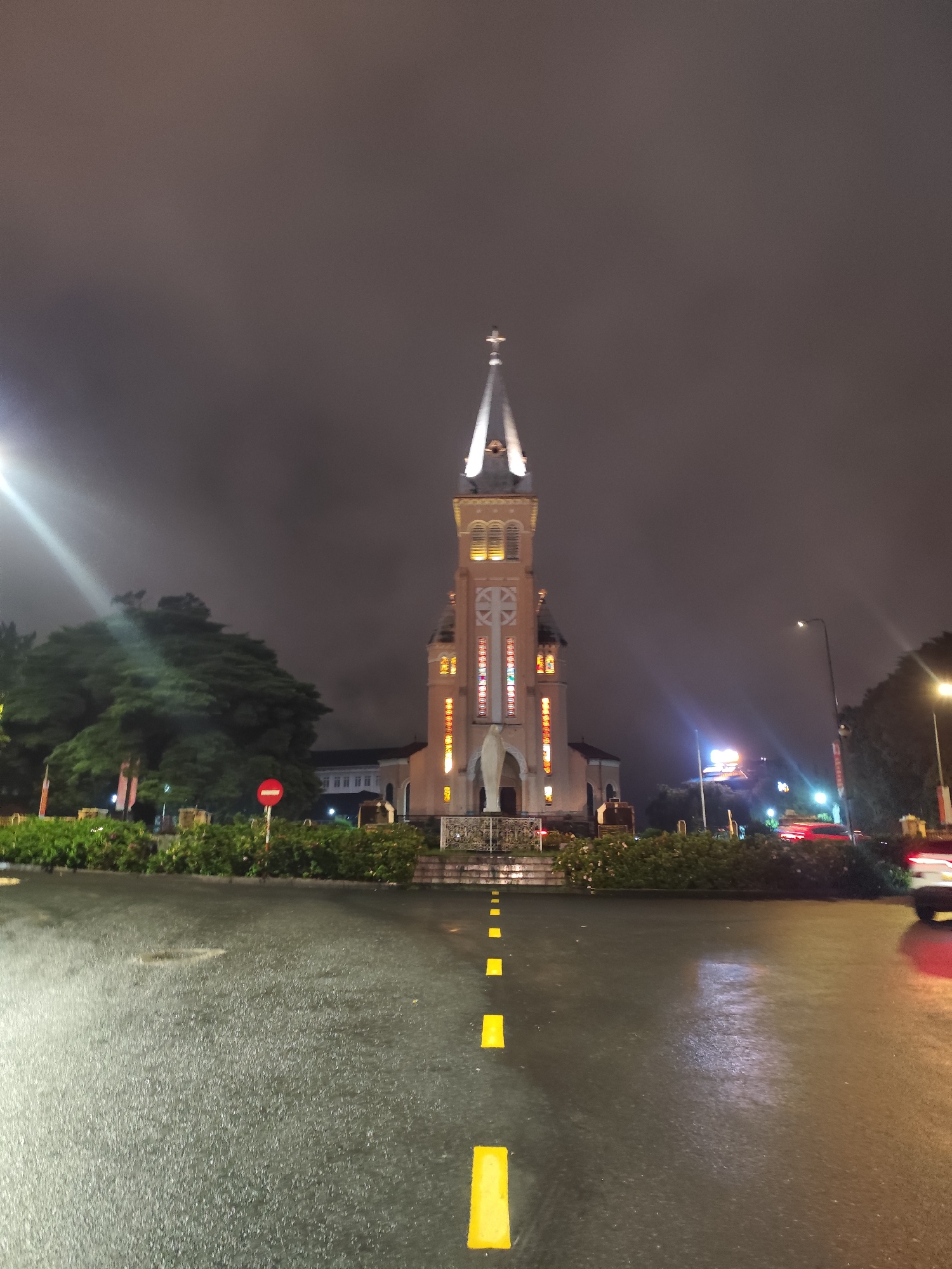 The image depicts a nighttime view of St. Nicholas Cathedral in Da Lat, Vietnam. The cathedral's illuminated tower and spire dominate the frame, set against a dark, cloudy sky. In the foreground, a wet asphalt road stretches towards the cathedral, marked with yellow rectangular lane dividers. Low-lying greenery lines both sides of the road, with a "Do Not Enter" sign visible on the left. Car headlights and other city lights provide illumination in the midground, suggesting vehicle traffic. The environment suggests a rainy or recently wet evening.